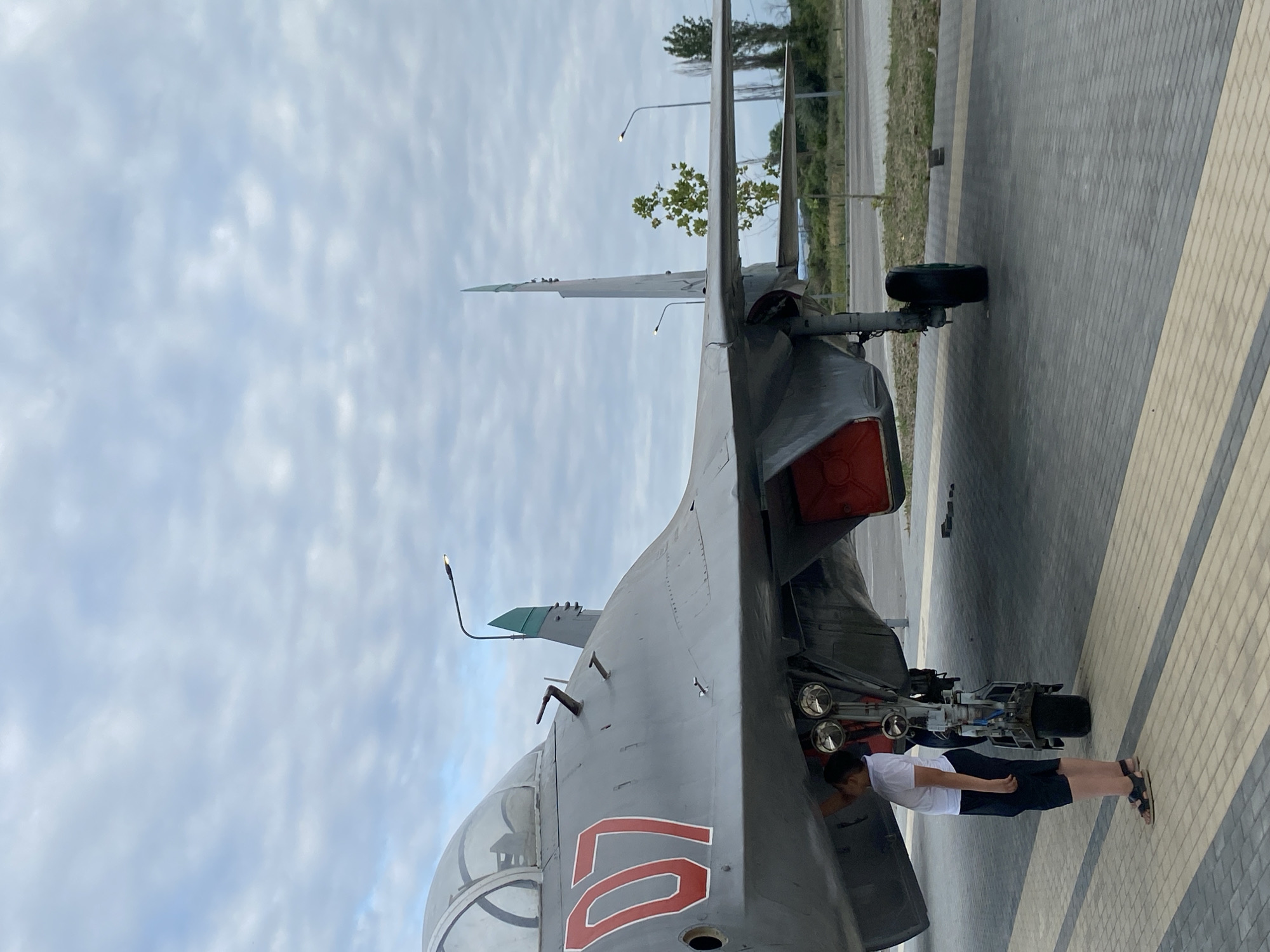 In Arablyar, Russia, a young person wearing a white t-shirt and dark shorts stands next to the landing gear of a grey military jet with the number "07" in red on its nose. The jet is parked on a paved surface with a patterned design of grey and yellow bricks. The sky is overcast with light grey clouds. In the background, there is a road, a grassy area with trees, and a few tall poles with lights. The scene suggests an outdoor display or museum exhibit.
