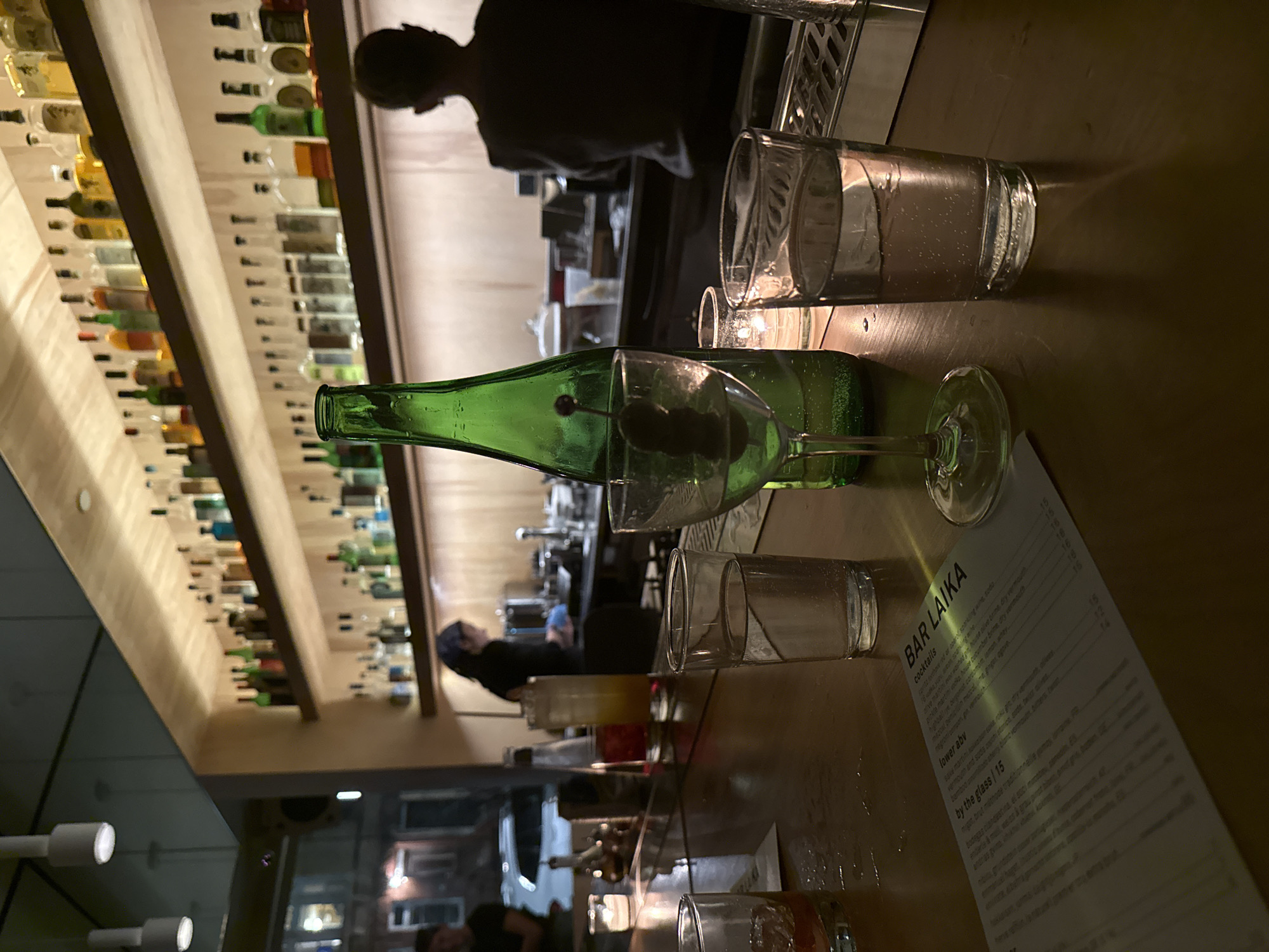 The image shows a bar scene at Bar Laika, a cocktail bar in New York City, United States. The scene features a green martini bottle with olives, glasses with liquid, and a menu. There are people in the background, including bartenders and patrons. The environment is dimly lit with a wooden bar and shelves of liquor bottles.