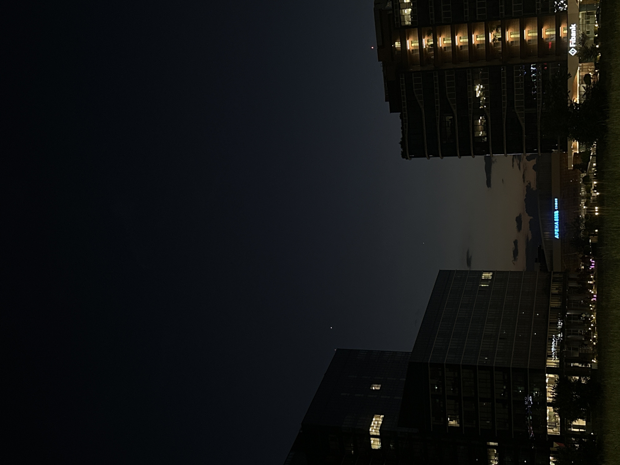 This is a night photograph of modern buildings in Sofia, Bulgaria. On the left, a large, dark glass building with illuminated windows is visible. To the right, a taller, multi-story building with a facade featuring rows of lit windows glows with warm light. In the background, a building with a sign that reads "ARENA" and "SOFIA" in neon blue is partially visible. At the base of the building on the right, a sign for "Fibank" is illuminated. The sky is dark, with a faint twilight glow near the horizon. The foreground shows a grassy area with some small lights. A single bright star or planet is visible in the upper sky. The overall scene depicts an urban environment at dusk or night.