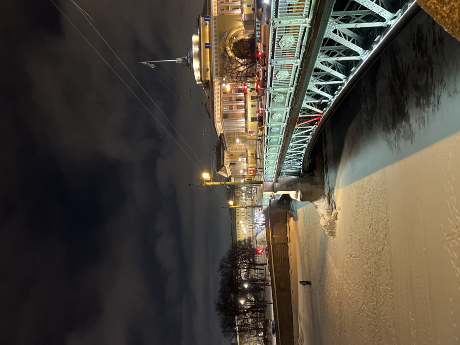 A nighttime perspective of a city bridge and buildings. The bridge, with its intricate metalwork, curves across the top. Below, a river is partially frozen with snow. Illuminated buildings line the riverbank, their yellow lights reflecting on the ice. Streetlights are visible, and bare trees stand along the embankment. The sky is dark and cloudy, enhancing the contrast with the bright lights of the city.
