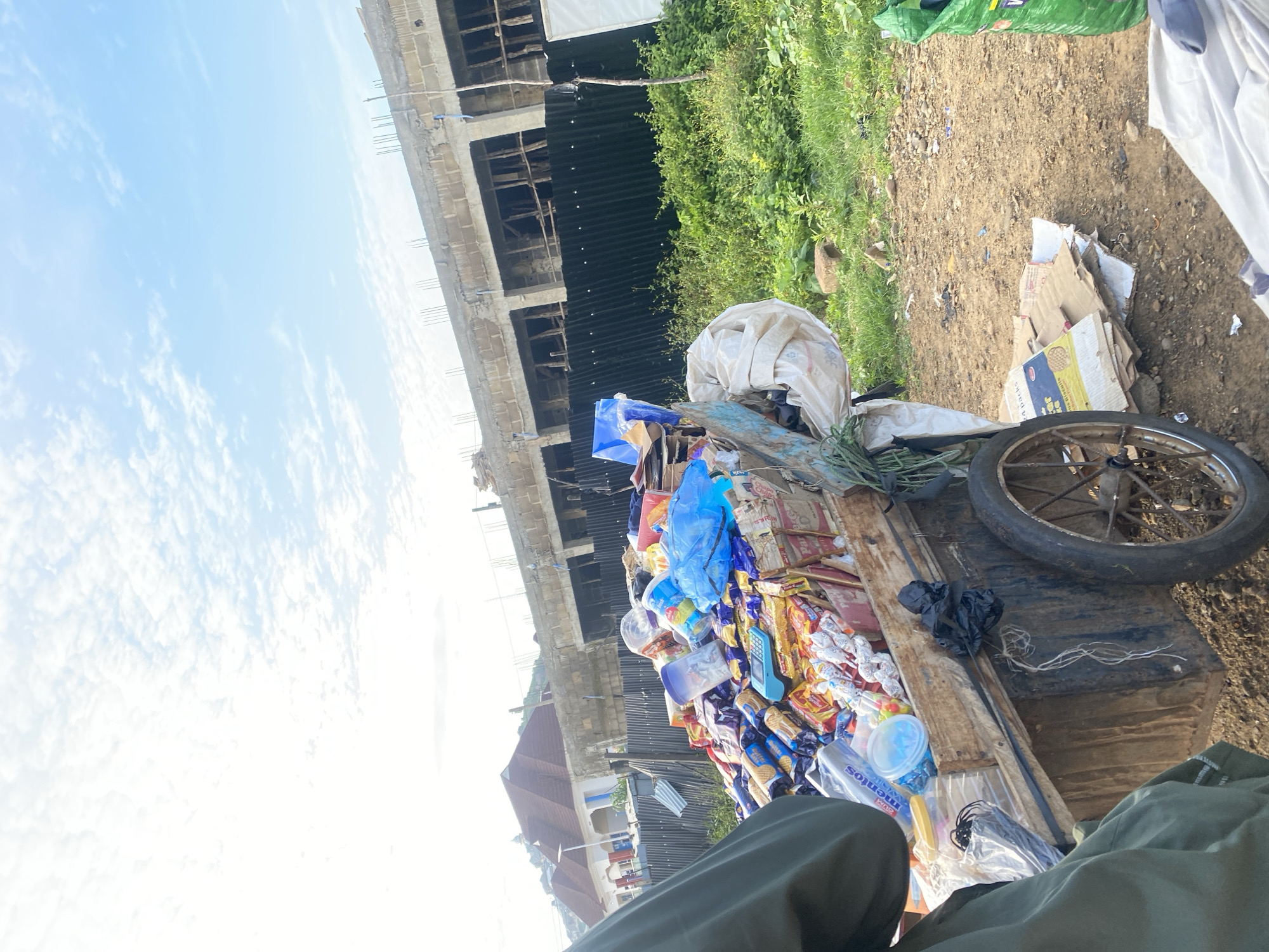 A rickety wooden cart filled with various packaged snacks and sweets is parked on a dirt road in Rafin Par, Nigeria. The cart has one large wheel and appears to be pulled by a person whose olive-green jacket is visible on the left side of the frame. Behind the cart, a black corrugated metal fence separates the street from a construction site with unfinished buildings. Lush green vegetation grows along the fence line. The sky is bright blue with scattered white clouds, and the sun is high, casting shadows. The ground is a mix of dirt and small stones, with some scattered cardboard boxes and debris near the cart.