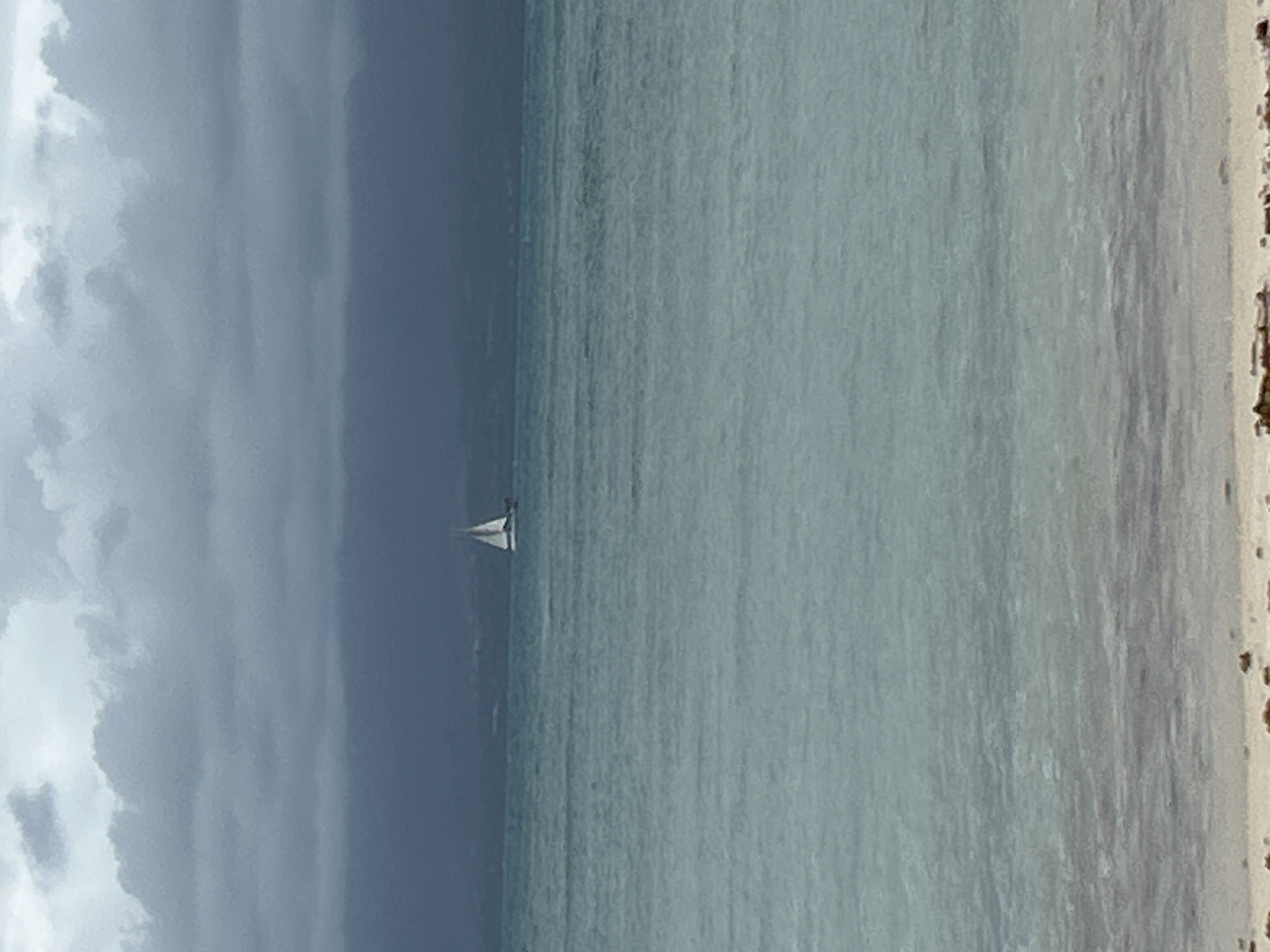 A horizontal landscape image shows a beach scene in Le Gosier, France. On the left side of the image, storm clouds dominate the sky above the ocean. The ocean itself is a gradient of deep gray-blue on the left to a lighter turquoise towards the beach on the right. A small white sailboat is located near the middle of the image. A sandy beach is visible along the right edge of the image.