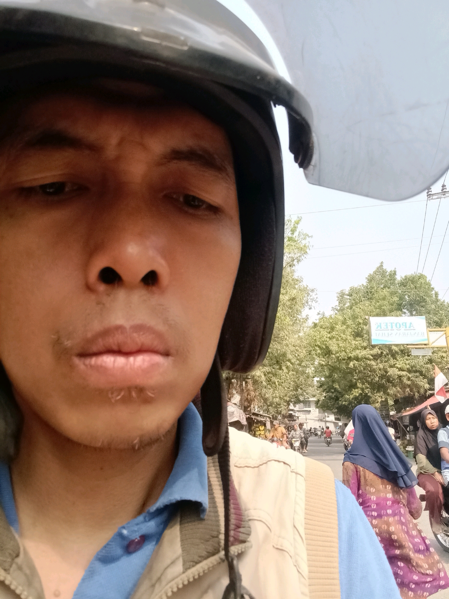 This is a selfie taken by a person wearing a helmet, likely on a motorcycle, on a street in Ujungrusi, Indonesia. The person's face is partially visible, with their eyes looking downwards. The background shows a street scene with trees, buildings, and other people on motorcycles. A sign with text in what appears to be Indonesian is visible on the right side of the frame. The overall atmosphere suggests a typical day in a bustling Indonesian town.