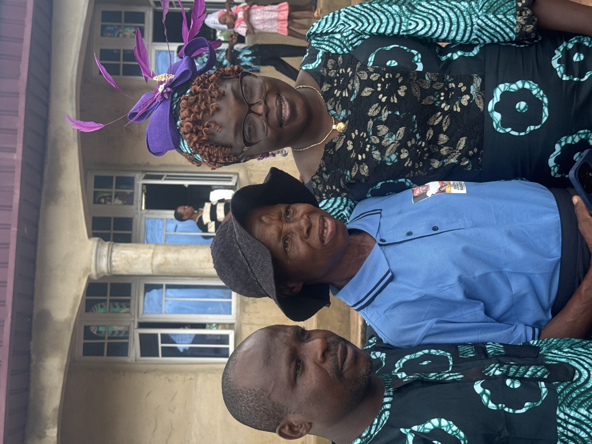 The image depicts three individuals posing for a photo outside a building.
The woman on the top left is wearing a black dress with a green and white patterned fabric, a purple hat with feather accents, and glasses. She is smiling. Below her is an older woman wearing a blue shirt and a dark-colored hat.
The man in the bottom center is wearing a black shirt with a green and white pattern that appears to match the dress of the woman above him. They are positioned in front of a building with a series of windows, some reflecting the sky.
Based on the prompt, the location is Eziudo, Nigeria.