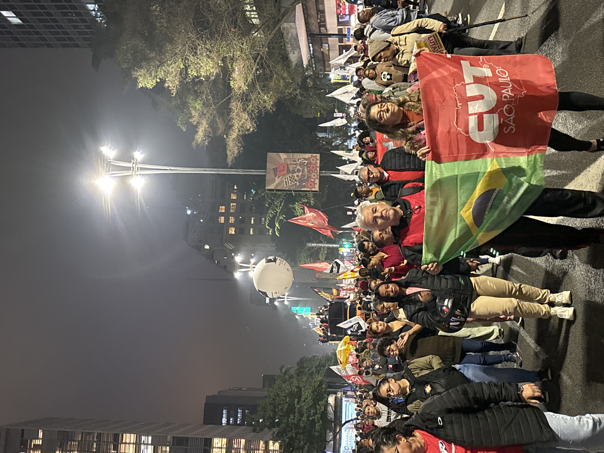 A nighttime crowd scene in São Paulo, Brazil, with people lining a street holding flags and banners, illuminated by streetlights. The Brazilian flag and a red banner reading "CUT SAO PAULO" are visible in the foreground. Buildings are seen on both sides of the street. The scene appears to be a protest or rally.
