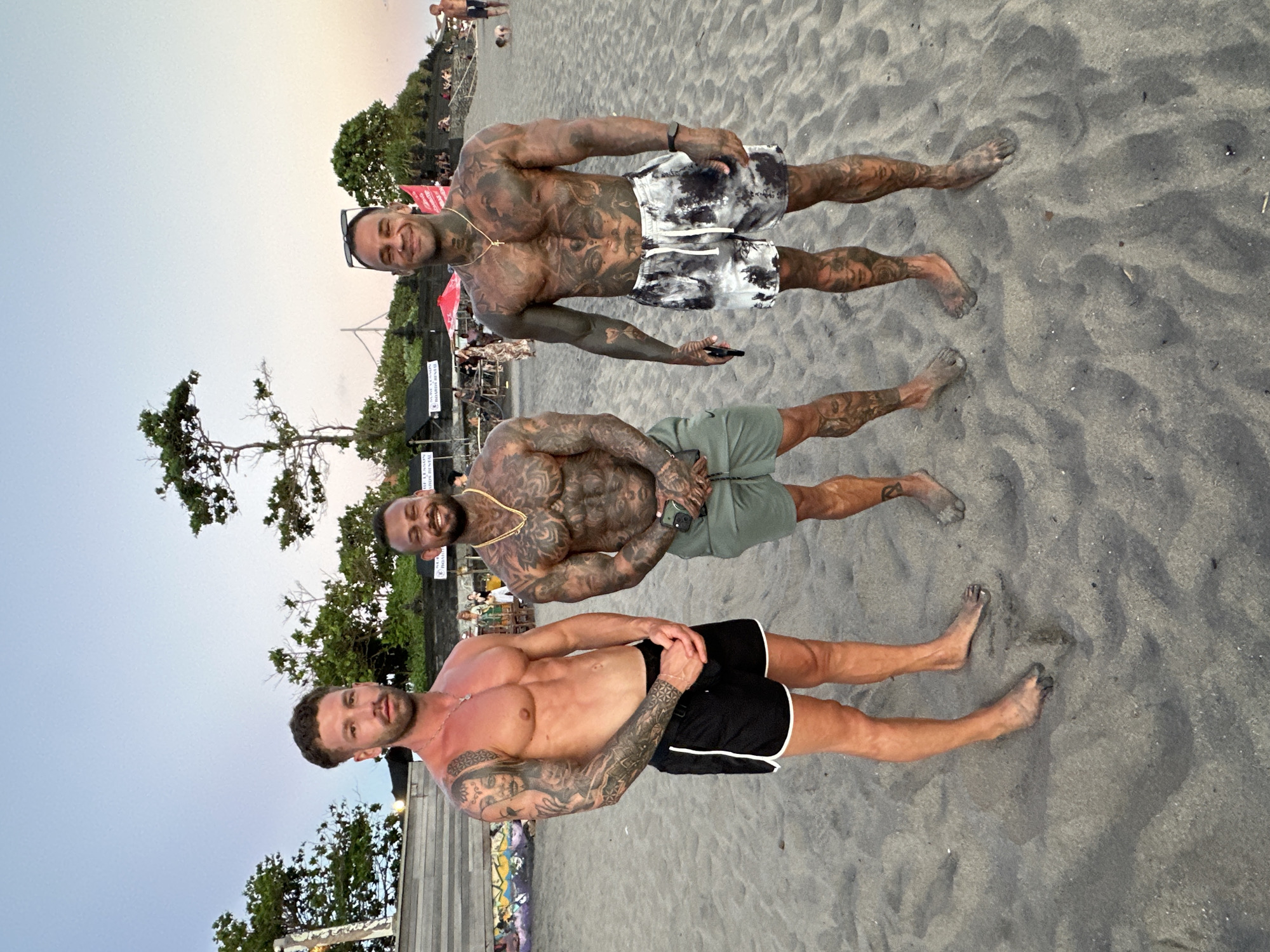 Three shirtless men with tattoos stand on a sandy beach, likely Canggu Beach in North Kuta, Indonesia. They are arranged vertically in the frame, posing for a photo. The man at the top wears sunglasses and black and white patterned shorts. The man in the middle wears a gold chain and green shorts. The man at the bottom wears black shorts with white trim. The beach appears to be near a wooded area with some structures visible in the background. The sky is overcast, suggesting either early morning or late afternoon.