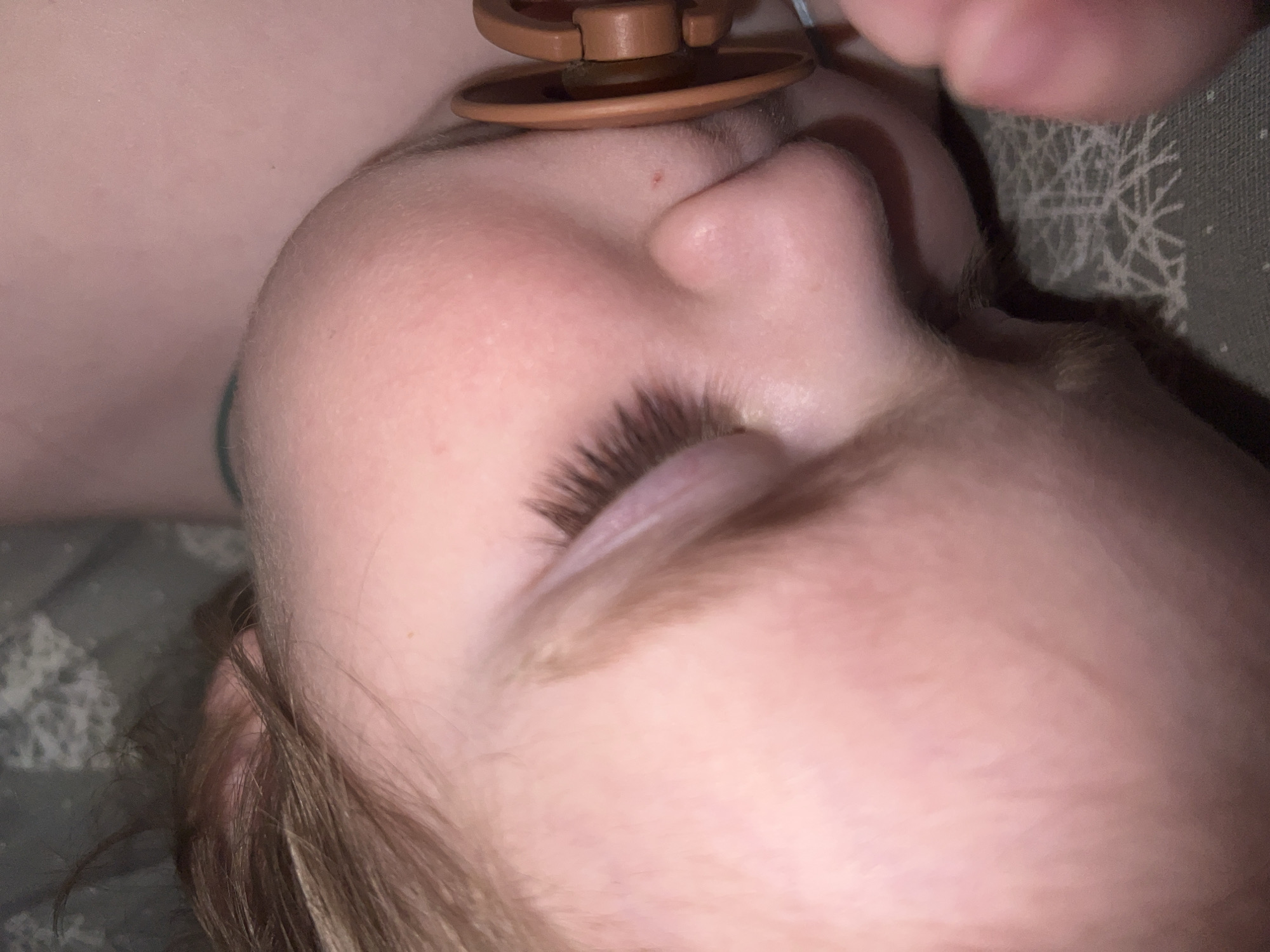A close-up, slightly out-of-focus image shows a child's face lying on a patterned fabric or bedding. A brown, disk-shaped object rests on the child's forehead, likely being held in place by a hand visible in the top right corner. The child's eye is closed, and their eyelashes are prominent. Strands of light brown hair are visible near the bottom of the frame. The image appears to be taken indoors. There is no information in the image that indicates the location of Noviy, Russia.