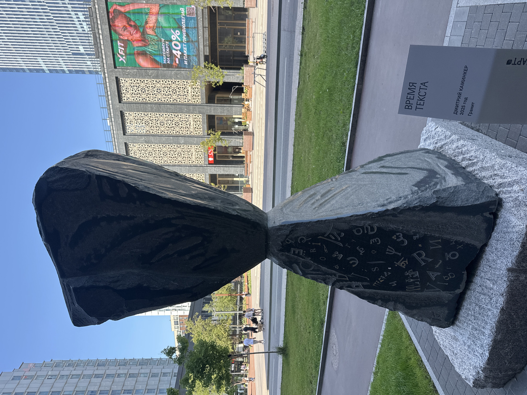 This is an outdoor photograph of a modern sculpture in an urban setting, likely in Moscow, Russia. The sculpture, made of dark stone, is shaped like an hourglass and is covered in various symbols and letters. It sits on a granite base in front of a modern building complex. In the background, there are tall buildings, some with reflective windows, and a billboard advertising a fitness center with a woman and the text "XFIT" and "-40%". There is a street in front of the building, with a sidewalk and a grassy area. Several people are walking in the background, including a couple with a stroller. The sky is clear and blue, indicating a sunny day. A sign next to the sculpture provides information about the artwork, including the title "ВРЕМЯ ТЕКСТА" (Time of Text), the artist "ДМИТРИЙ КАМИНКЕР" (Dmitry Kaminker), the year "2025 год" (2025), and the material "Гранит" (Granite).
