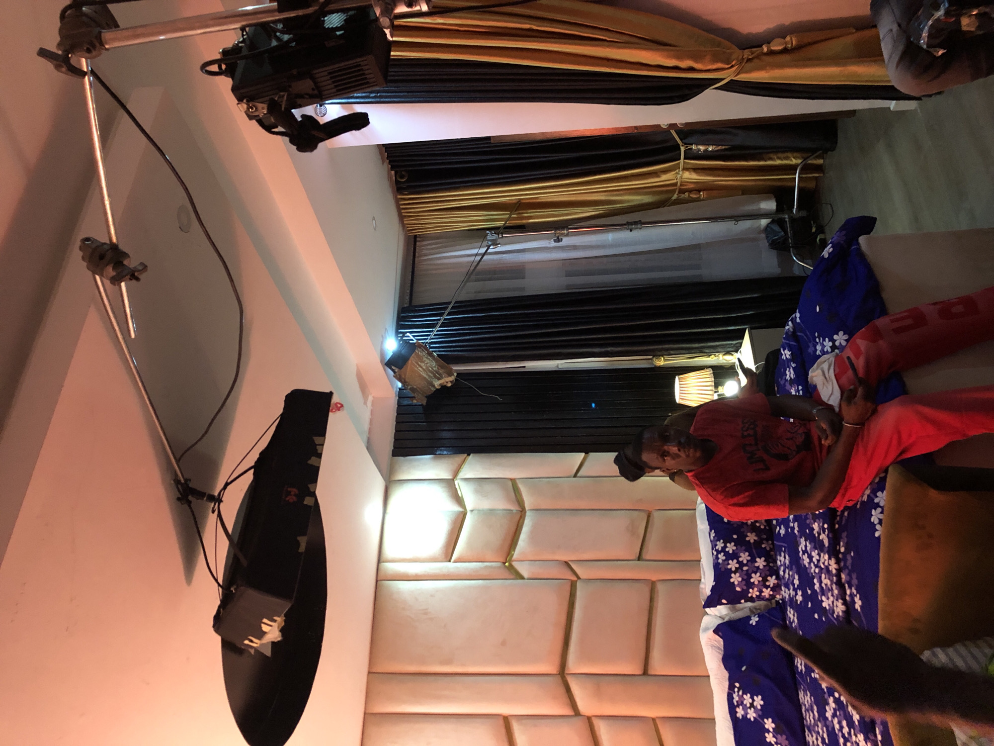 An indoor scene, rotated counter-clockwise, depicts what appears to be a bedroom or hotel room set up for filming or photography. A male subject, wearing a red t-shirt with the word "LIMITLESS" visible in white capital letters and red pants, lies on a bed. He has a dark cap on his head and looks towards the camera. His left hand is raised, possibly making a "V" sign.
The bed is covered with a blue duvet featuring a white floral pattern. The headboard is upholstered in a light-colored material with a geometric, padded design, some sections outlined in gold. The room's walls are white.
Extensive professional lighting and camera equipment are visible, indicating a production setting. A long metal pole extends across the ceiling, supporting a large black light fixture. Another bright light source is visible on a stand or mounted, and cables run connecting the equipment. A large, black, semi-circular object, possibly a reflector or another light, is mounted on a wall.
In the background, black and gold curtains are drawn, with white sheer curtains behind them. A bedside lamp with a woven shade is visible near the person's head. The artificial lighting gives the scene a warm glow. Another hand is partially visible in the bottom right corner of the image.