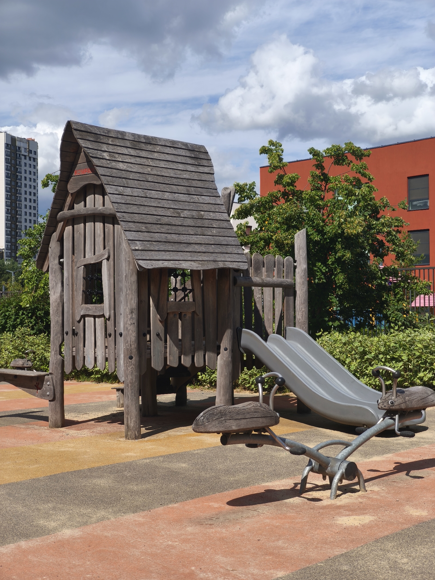 An outdoor playground is depicted, featuring a rustic wooden playhouse with a peaked roof, a gray slide, and a seesaw. The playhouse has a small window and is constructed from weathered wood planks. Adjacent to the playhouse is a bright orange building, and in the background, a tall, modern apartment building stands against a sky with scattered clouds. The ground is surfaced with a multi-colored, textured material, likely rubberized safety surfacing, with distinct orange, tan, and gray sections. Green bushes and trees surround the play area. The scene is set in Moscow, Russia.