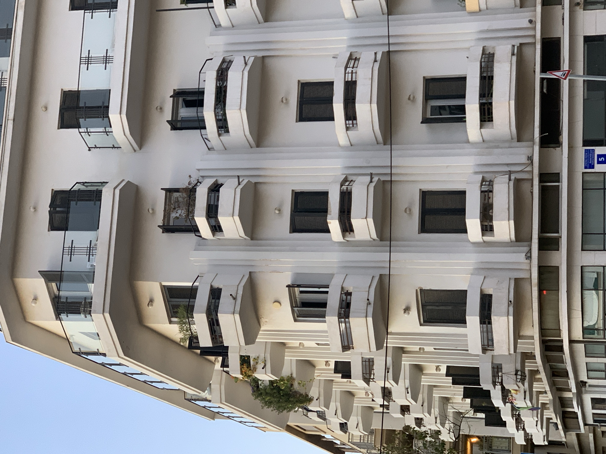 The image shows the facade of a building in Tel Aviv, Israel, characterized by a white exterior, repetitive geometric patterns, and protruding balconies. The architecture is reminiscent of Bauhaus or International Style, common in Tel Aviv. The building has a series of windows and balconies with dark shutters. Street signs and architectural details of neighboring buildings are visible. The sky is light blue.