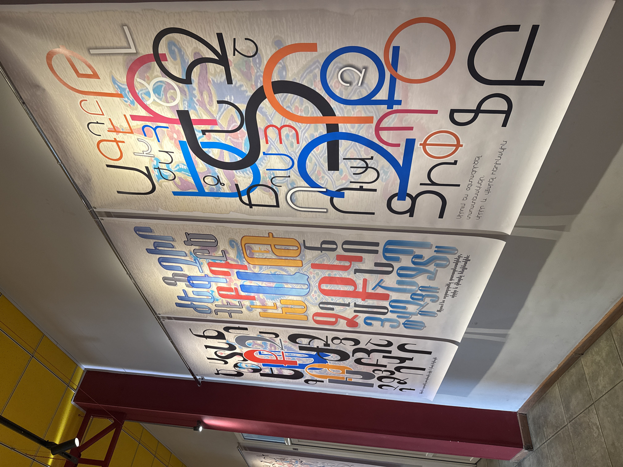 The image captures an installation in what appears to be a building in Yerevan, Armenia. Two large posters are suspended from the ceiling, displaying a variety of stylized Armenian letters in different colors and sizes. Below the letters, there is Armenian text. The first poster shows the letters layered and superimposed on each other, while the second poster shows them arranged in a more grid-like fashion. The ceiling above the posters is white, while the walls are painted in yellow and gray. A red beam structure is visible on the lower left side.