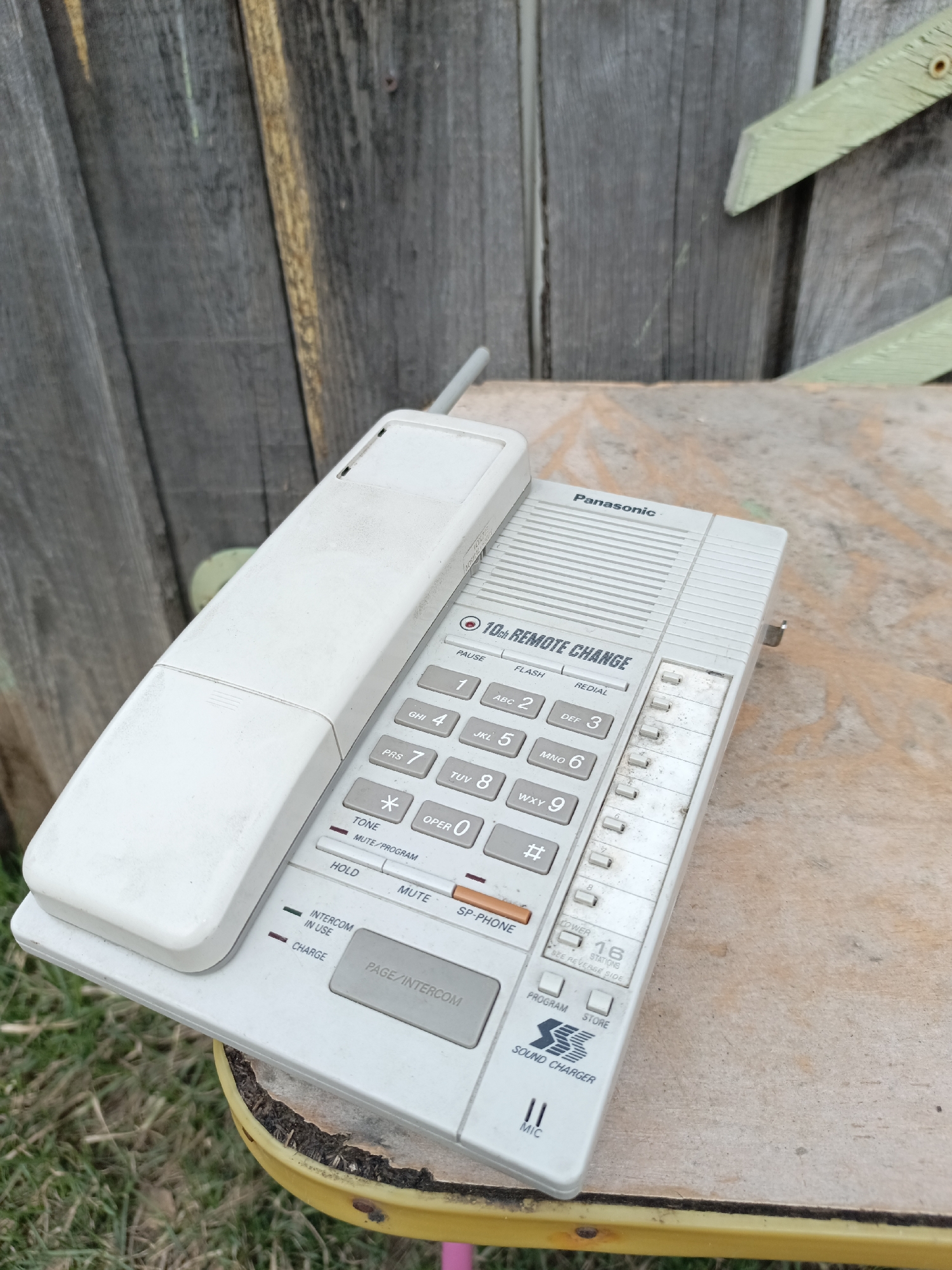 An old, off-white Panasonic cordless telephone base station and its matching handset are prominently featured, resting on a weathered, light-colored wooden surface. The phone components exhibit signs of age, including dust, dirt, and some discoloration. The base station has a full numeric keypad (1-9, *, 0, #) with associated letters (ABC, DEF, GHI, JKL, MNO, PRS, TUV, WXY), alongside function buttons such as "PAUSE," "FLASH," "REDIAL," "TONE," "MUTE/PROGRAM," "HOLD," "MUTE," and "SP-PHONE." Indicator lights are visible for "INTERCOM IN USE" and "CHARGE." Below the keypad, buttons for "PAGE/INTERCOM," "PROGRAM," and "STORE" are present. Text on the device also includes "Panasonic," "10ch REMOTE CHANGE," "POWER," "16 STATIONS," "SEE REVERSE SIDE," "SOUND CHARGER," and "MIC."
The setting is outdoors, with a dark, weathered wooden fence forming the background. A light green wooden element, possibly a decorative piece or part of the fence, is visible in the upper right. Beneath the wooden surface the phone rests on, hints of green grass and a yellow object (possibly a furniture leg) can be seen. The lighting suggests it is daytime. No people or specific activities are depicted. The location context for this image is Chaikovsky, Russia.