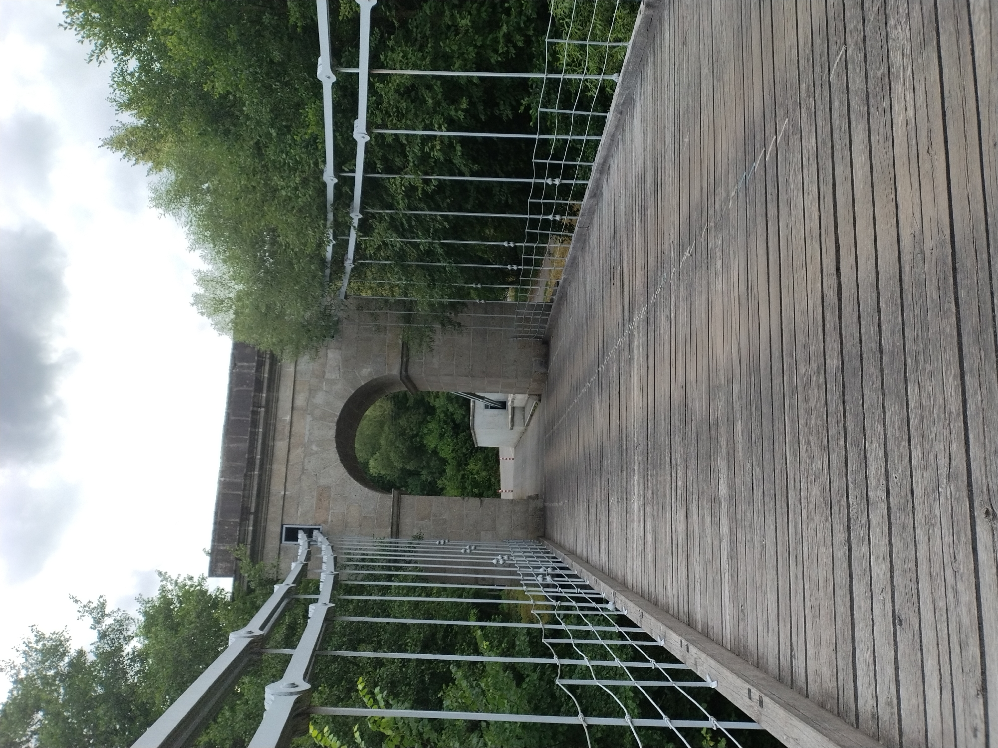 The image shows a perspective view of a wooden suspension bridge in Stádlec, Czechia. The bridge has a weathered wooden deck and white metal railings supported by vertical posts. Beyond the railings, dense green trees line both sides of the bridge. In the distance, an arched stone structure forms an entrance or gateway. The sky is overcast with heavy clouds. The scene is static, with no people or actions visible.