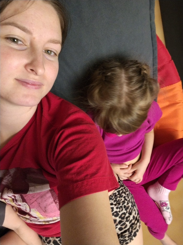 The image captures a close-up, high-angle selfie of a woman with a young child resting beside her, likely indoors on a bed or sofa.
The woman, positioned on the left side of the frame, has light skin, light brown hair pulled back, and green eyes. She is looking directly at the camera with a slight, gentle smile. She is wearing a red short-sleeved t-shirt, which appears to have a design featuring a cartoon character (possibly Mickey Mouse or a similar figure) in shades of pink, white, and brown on the lower left side. Below her shirt, a portion of her lower body is visible, dressed in leopard print shorts or underwear. Her right arm is visible, positioned lower in the frame.
To the right of the woman and slightly behind her is a young child, whose head and upper body are mostly out of focus and turned away from the camera, facing downwards. The child has light brown hair styled with two small braids or buns on either side of a central parting. She is wearing a bright fuchsia or magenta short-sleeved t-shirt and matching fuchsia pants. Her hands are clasped together in her lap. Her feet, visible at the bottom right, are wearing light pink socks with a white cartoon character design (possibly Hello Kitty) on them.
The background consists of soft furnishings. Immediately behind the woman's head is a dark gray cushion or pillow. To the right, beneath and behind the child, there is a colorful cushion or bedding with broad stripes of orange and red. The overall impression is a relaxed, intimate, and casual moment shared between the woman and the child. The lighting suggests an indoor setting during the daytime. No text is visible in the image.