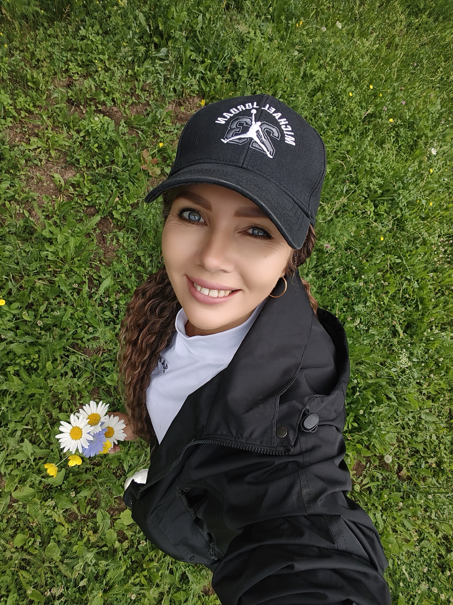 A woman is pictured in a selfie taken outdoors on a grassy field in Noviy, Russia. She is wearing a black baseball cap with the iconic "Jumpman" logo and text that appears to read "MICHAEL JORDAN" and "TORONTO". She also has on a white shirt and a black jacket. Her blue eyes are captivating, and she is smiling with her teeth showing. Her curly brown hair falls over her shoulder. In her left hand, she holds a small bouquet of white daisies and a single blue flower, with a few yellow wildflowers scattered nearby in the grass. The lighting suggests it is daytime, and the lush green grass indicates a mild climate. The woman's expression and the presence of flowers suggest a relaxed and pleasant mood.
