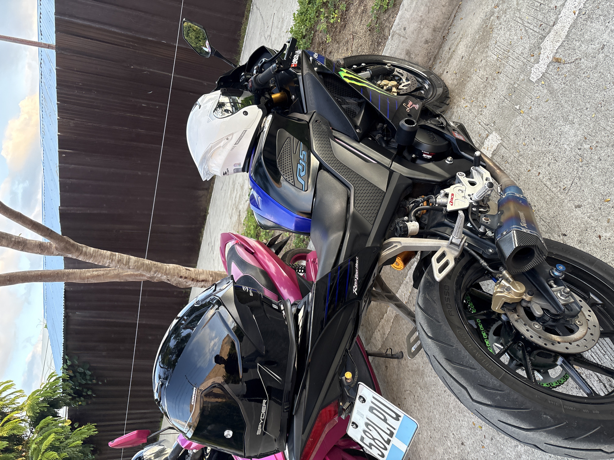Two motorcycles are parked on a concrete surface in what appears to be a residential area in Davao City, Philippines. The motorcycle in the foreground is a black Yamaha R15, identifiable by the "R15" graphics on its fuel tank and the "YAMAHA" logo on its fairing. It is equipped with a black exhaust pipe with a carbon fiber tip and green chain. A white helmet with a clear visor, brand marked as "SPYDER," is placed on the seat. In the background, another motorcycle, possibly a scooter, is visible with a pink seat and frame, and a pink passenger grab rail. A black helmet, also brand marked as "SPYDER," rests on its seat. The motorcycles are parked in front of a brown wooden fence topped with a blue corrugated metal roof. Green foliage from trees is visible overhead and to the left. The ground is a concrete pavement with some fallen leaves. The sky is partly cloudy with hints of sunset colors. A license plate with the text "5821 PU" is attached to the rear of the black motorcycle.
