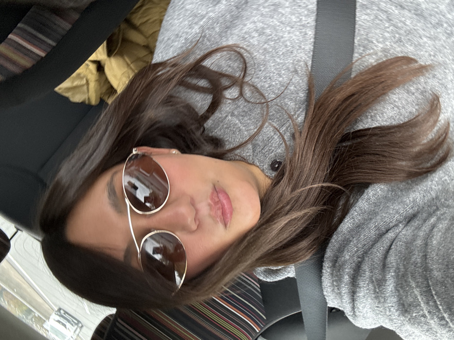 A female subject is depicted in a medium-shot, from the chest up, seated within a vehicle in Vidauban, France. She has long, dark brown hair parted on the left, extending over her shoulders and chest. She wears gold-rimmed, oval-shaped sunglasses with reflective brown lenses, which show distorted reflections of what appear to be building facades. Her lips are slightly pursed, and a small, light-colored stud earring is visible in her right ear. She is dressed in a gray, textured long-sleeved top or sweater with a dark button visible on the front near her collarbone. A dark-colored seatbelt diagonally crosses her chest. The vehicle's interior behind her features a dark seat with a multi-colored striped fabric pattern (dark red, brown, black, and white lines) visible on the left. To the right, behind her, a crumpled tan or khaki-colored garment or blanket is visible on the seat. The subject is looking directly towards the camera.