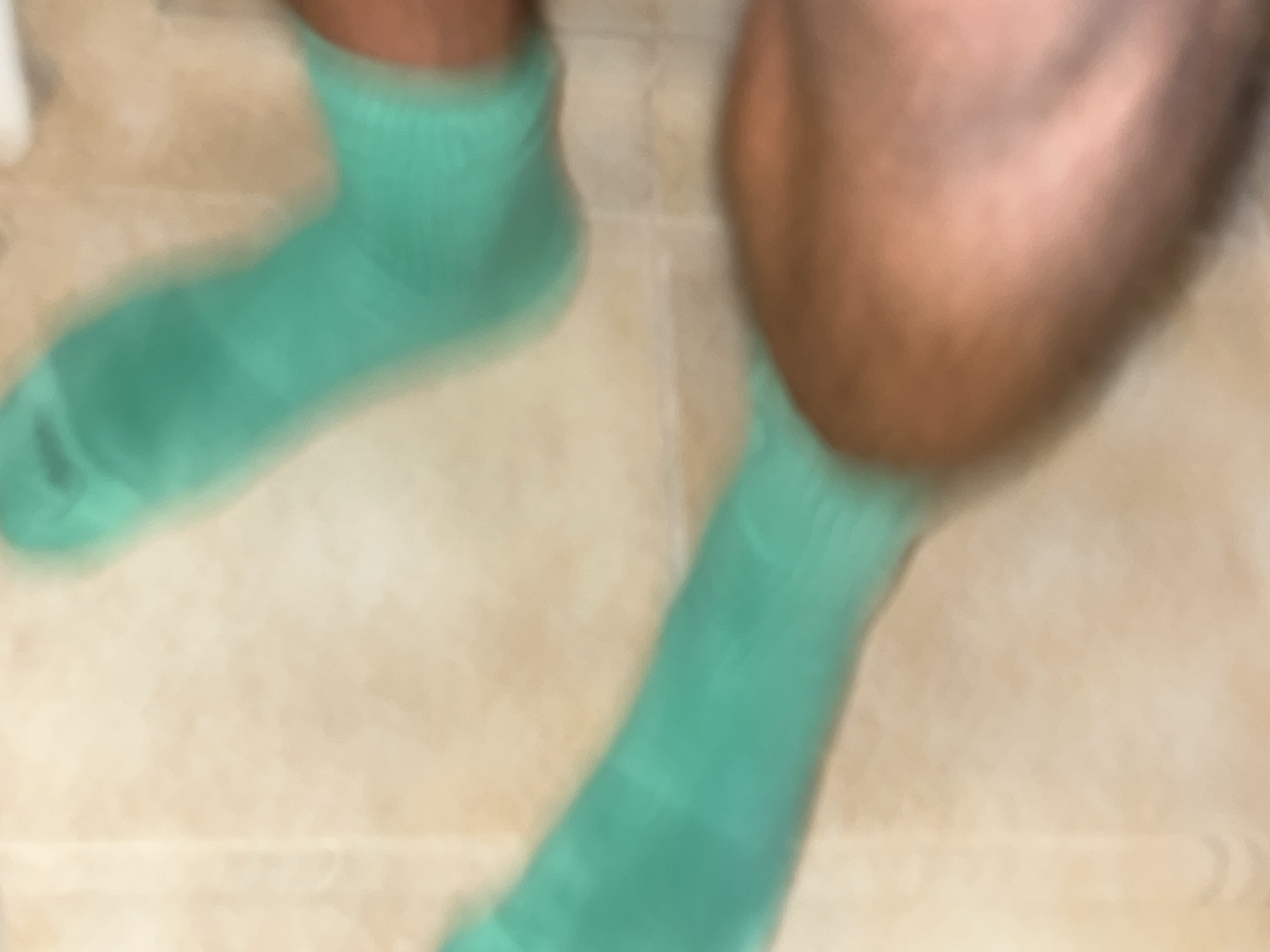 The image is a blurry, close-up shot of a person's legs wearing bright green socks. The background appears to be a tiled floor, possibly in a bathroom or kitchen. The lighting is soft and diffused. The composition is abstract due to the blurriness, focusing on the textures and colors of the socks and skin. There is no discernible indication of the location being the city of Budva, Montenegro.
