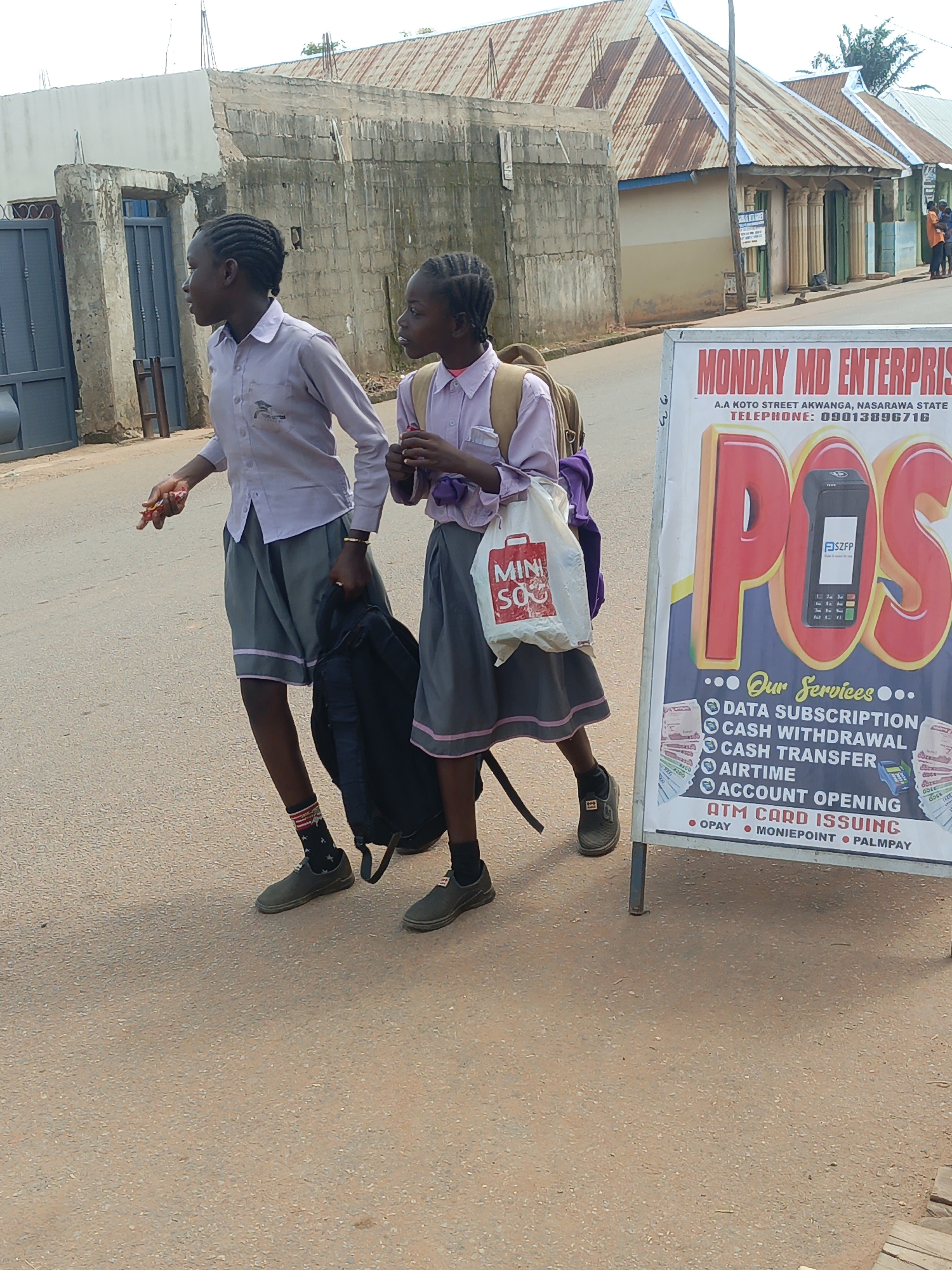 Two young girls, dressed in school uniforms consisting of light purple shirts and gray skirts, walk side-by-side on a paved street. They are likely in Akwanga, Nigeria, as indicated by the "MONDAY MD ENTERPRISE" sign visible in the foreground. The sign advertises various financial services, including "DATA SUBSCRIPTION," "CASH WITHDRAWAL," "CASH TRANSFER," "AIRTIME," and "ACCOUNT OPENING," listing "OPAY," "MONIEPOINT," and "PALMPAY" as supported platforms. The girl on the left, closer to the viewer, wears black socks with a red and white pattern and dark shoes. She carries a black backpack in her right hand and holds a piece of candy or food item in her left hand. The girl on the right carries a white bag with red lettering that reads "MINI SOS" and a brown backpack strapped to her shoulders. Behind them, buildings with corrugated iron roofs line the street, and a concrete wall with a blue gate is also visible. The overall scene depicts an everyday street view in a Nigerian city.