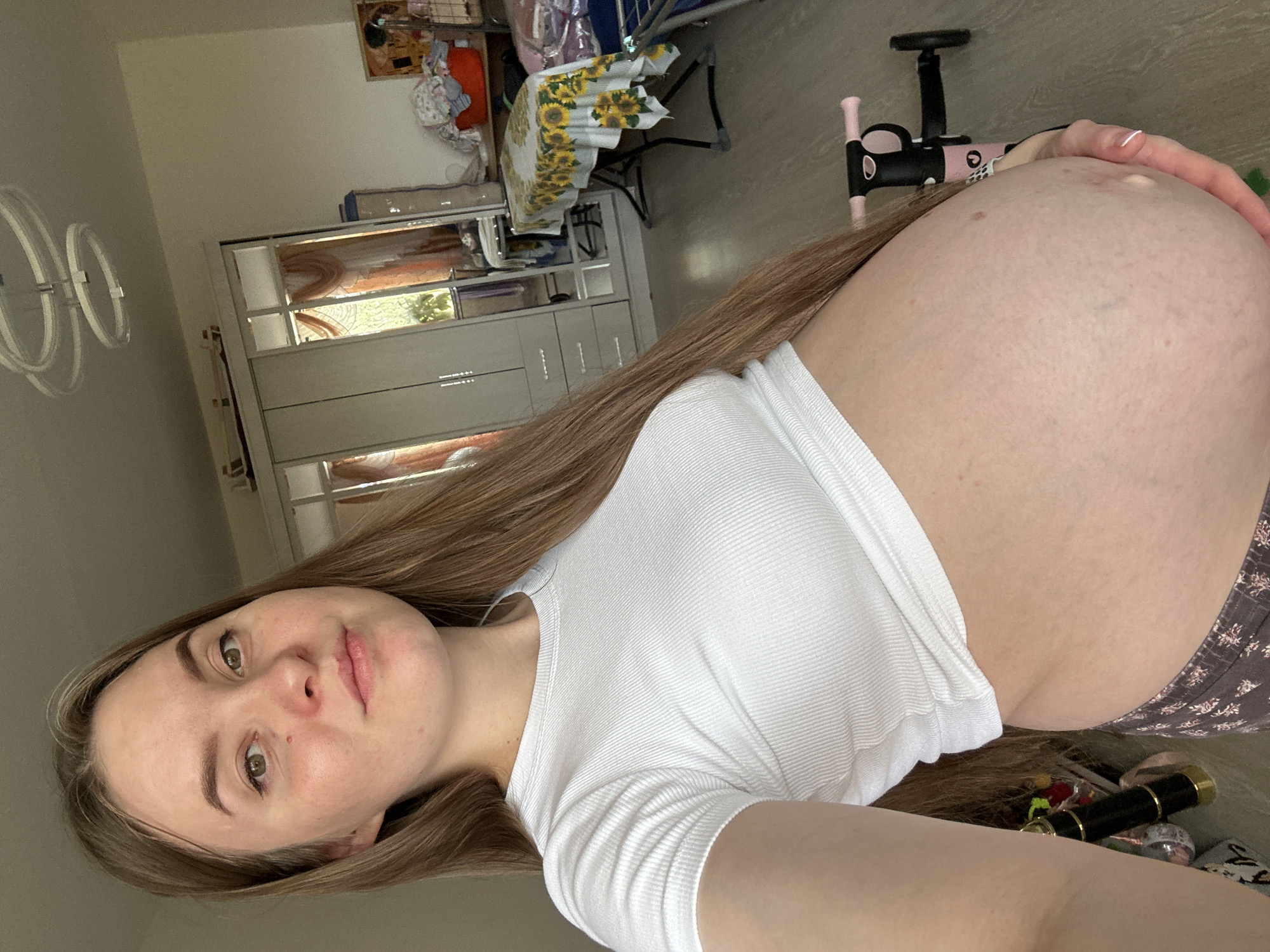 The image shows a pregnant person in a room that appears to be a bedroom or living space, likely in Noviy, Russia. The person is standing and facing the camera, with their belly prominently displayed. They are wearing a white, ribbed crop top and patterned pajama bottoms. The background includes a large wardrobe with mirrored doors, a table with a sunflower-patterned tablecloth, and a laundry basket filled with clothes. A ceiling light fixture with concentric rings is visible overhead. A child's pink and black scooter is positioned behind the person. The lighting suggests it is daytime.