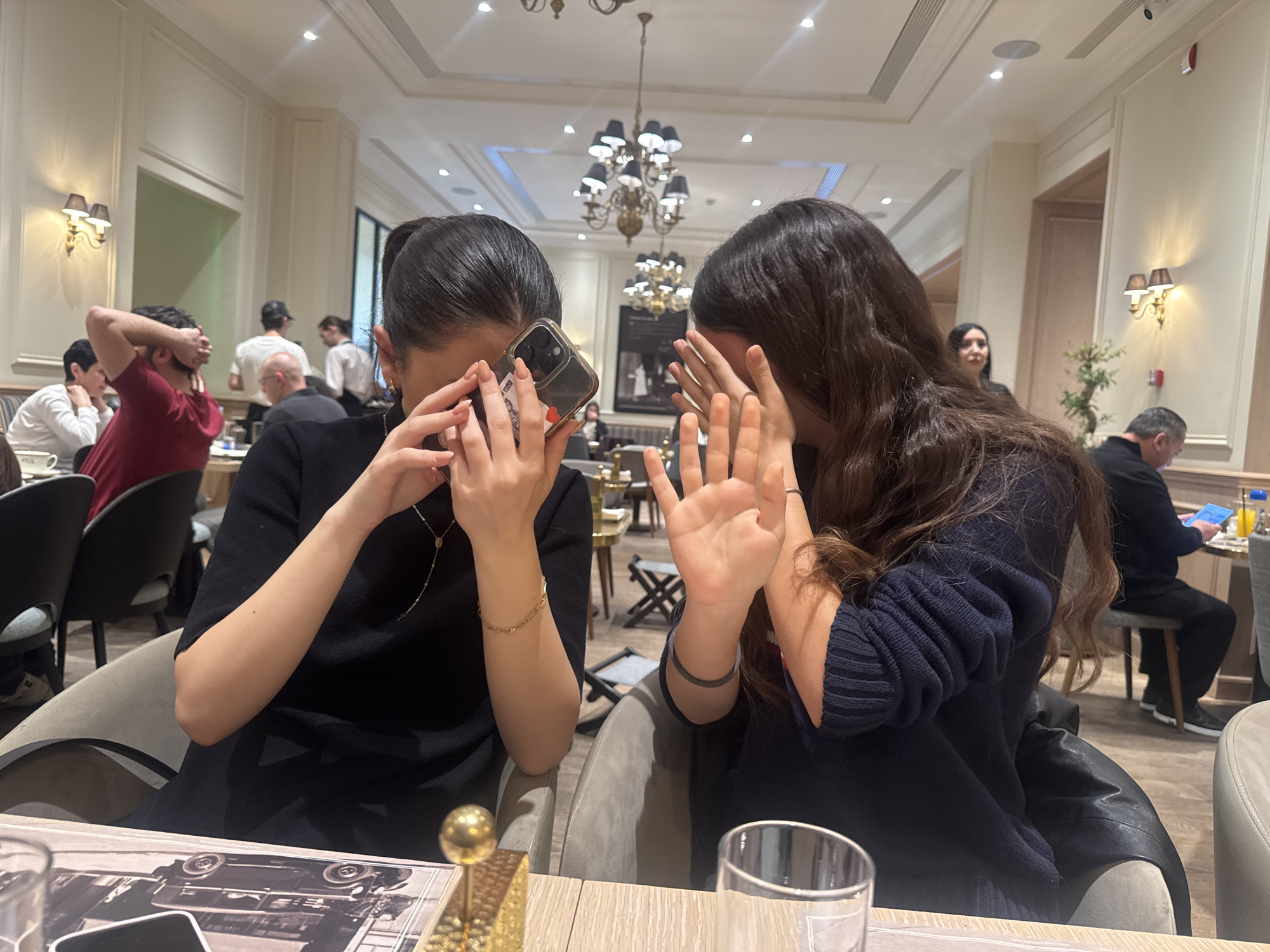 This indoor photograph captures a scene within an upscale restaurant or cafe in Yerevan, Armenia, likely in the evening. In the immediate foreground, two young women are seated at a table, both playfully or shyly covering their faces from the camera. The woman on the left, with dark hair pulled back, wears a black top, a gold chain necklace, and a gold bracelet. She holds a smartphone with a clear case and a visible red heart design on its back, using it to partially obscure her face. Her nails are manicured. The woman on the right, with long dark wavy hair and a dark blue ribbed sweater, covers her face with her hands, revealing a ring on her right hand.
The background shows a softly lit, elegant dining environment with multiple ornate chandeliers hanging from the ceiling and decorative wall sconces providing additional light. Several other people are visible: a man in a red shirt on the left, numerous blurred staff members in white shirts and dark attire, and other patrons dining or conversing. One man in the far background is seen looking at a tablet with a bright blue screen. The decor includes sophisticated wall paneling, a large framed picture on a distant wall, and a small plant. On the table in the foreground, part of a menu or placemat featuring a black and white image of an old car, a golden decorative item, and a clear drinking glass are visible. The atmosphere suggests a refined yet active restaurant setting.