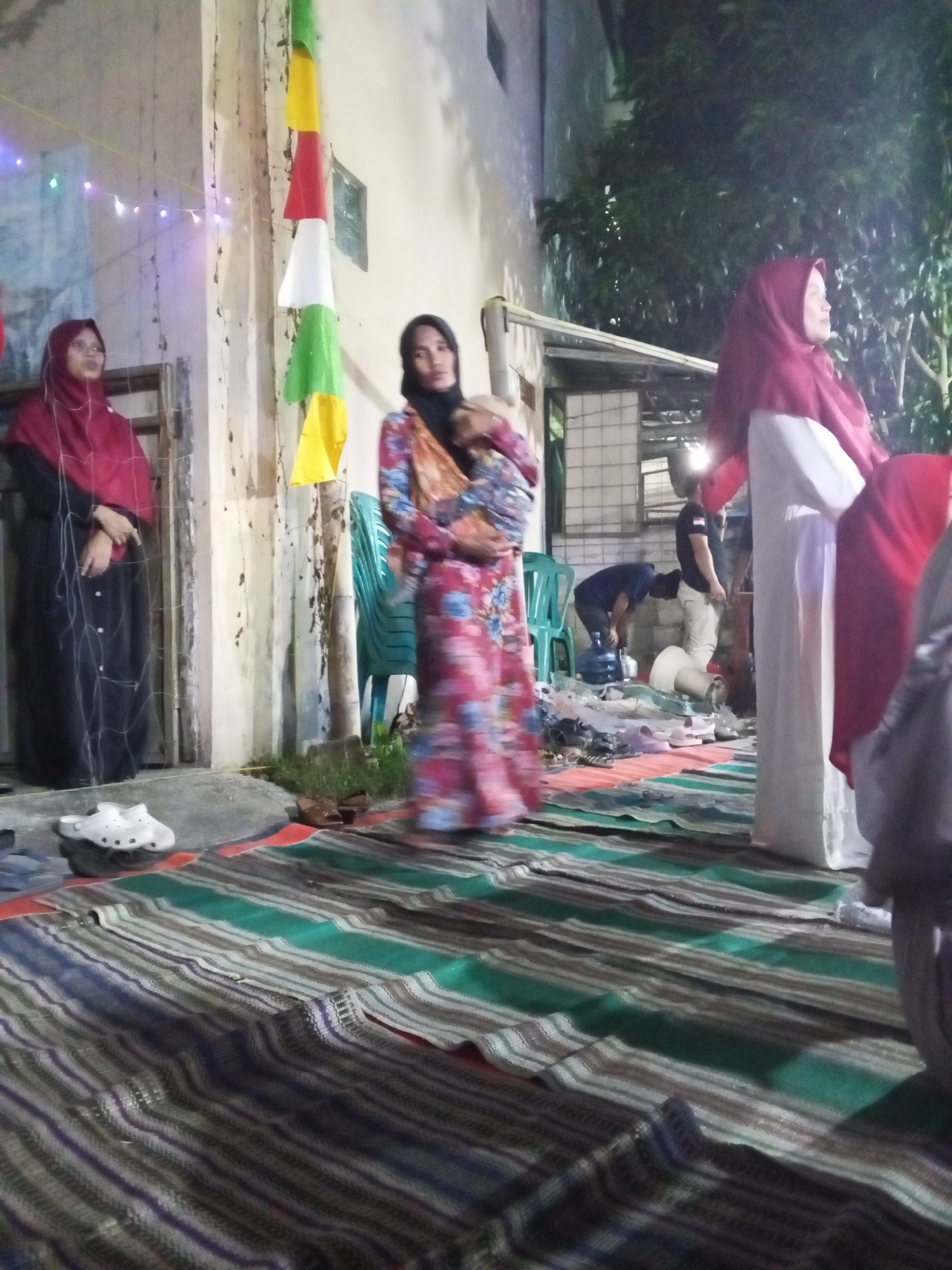 The image depicts an outdoor evening gathering in Ciruas, Indonesia, with a focus on a group of people. The scene is illuminated by artificial lights, some of which are string lights with colored bulbs. In the foreground, a patterned mat with horizontal stripes in green, brown, and white covers the ground. To the left, a woman in a red hijab and a black abaya stands near a doorway. Further back, a woman in a floral-patterned dress and a black hijab holds a baby. Behind her, a man in a black t-shirt and light-colored pants is bending over a large blue water jug. Several green plastic chairs are visible in the middle ground. On the right side of the image, a woman in a red hijab and a white garment stands, and another person in a red hijab is partially visible. A white object, possibly a roll of fabric or paper, is on the ground. The background shows a building with a tiled roof and some trees. The overall atmosphere suggests a communal event, possibly a celebration or a social gathering.
