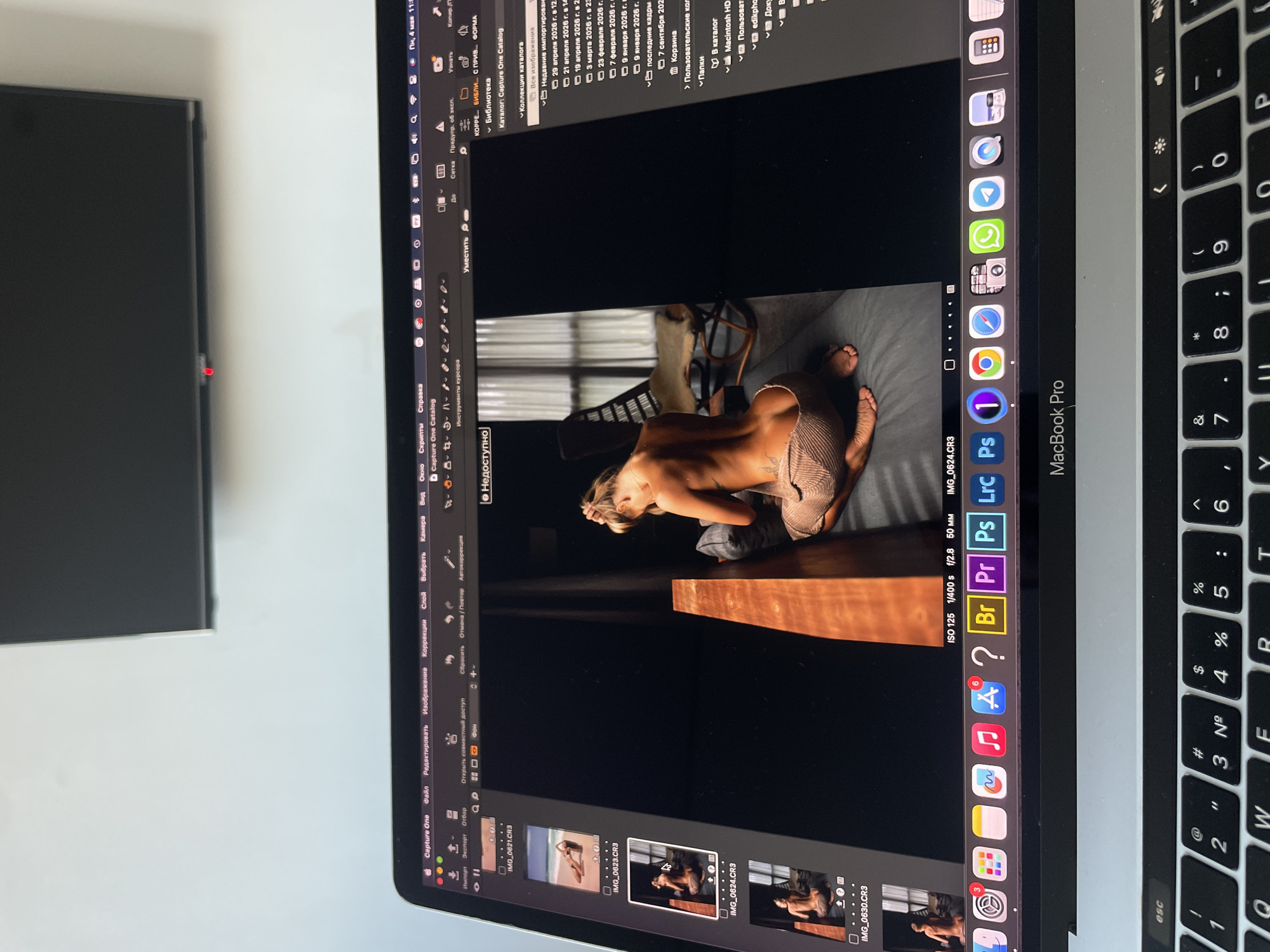 The image depicts a screen capture of a MacBook Pro displaying the Capture One software interface, likely being used to edit a photograph of a lightly-dressed woman posing in what appears to be an interior setting. The software interface includes various image editing tools and a thumbnail gallery of images. The MacBook Pro is positioned on a white surface, and a black monitor is partially visible to the left of the MacBook. The keyboard of the MacBook Pro is visible on the right. The location in the city of Kazan, Russia is not explicitly determined by the image.