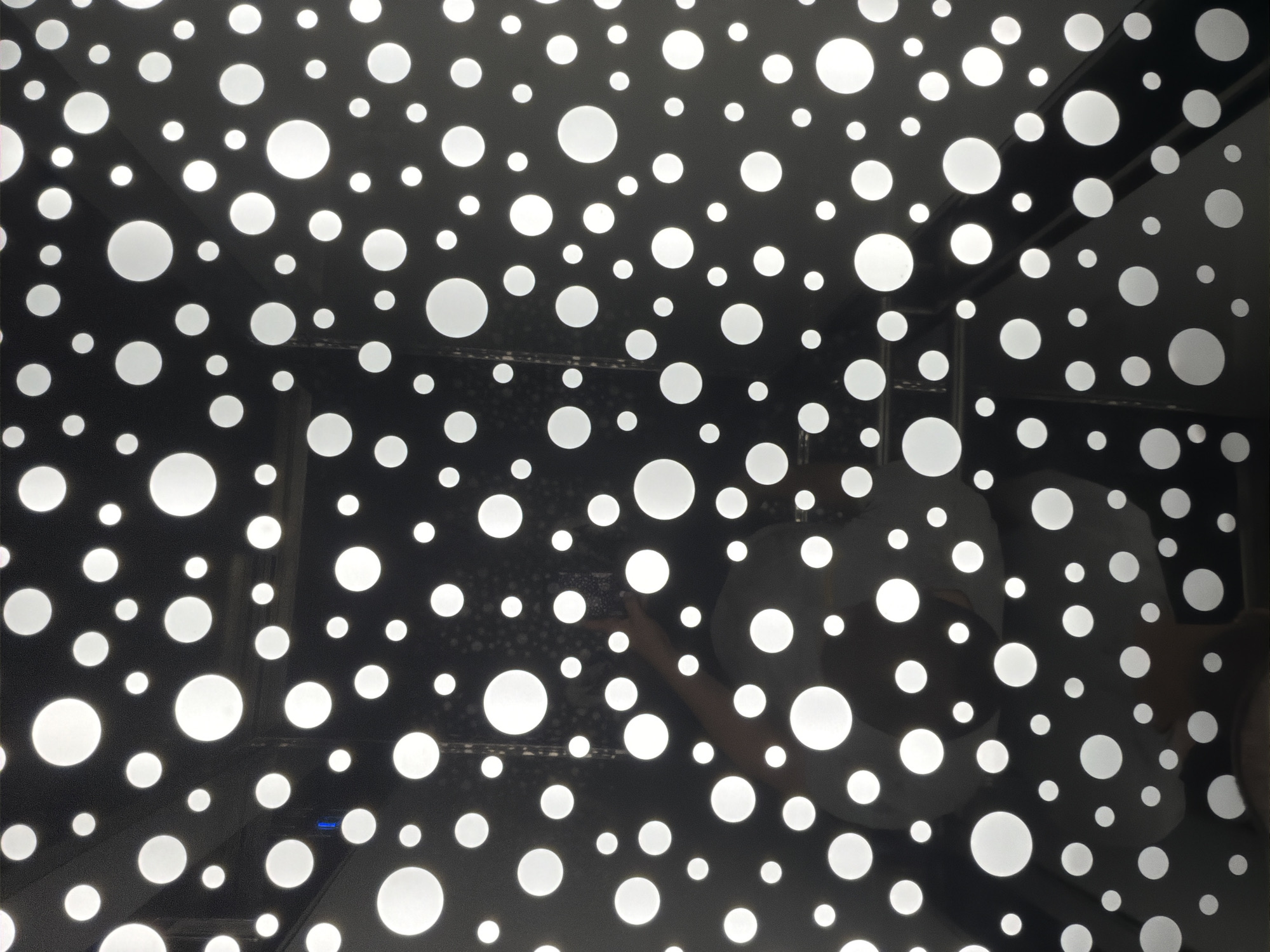 The image shows a black surface with numerous white polka dots of varying sizes. A person is visible reflected in the surface, taking the photo with their hand and phone. The dots appear to be illuminated from behind, creating a backlit effect. The setting is an indoor environment, possibly a building with a modern design. There are no specific indicators in the image to pinpoint its location within Dolno Ablanovo, Bulgaria.