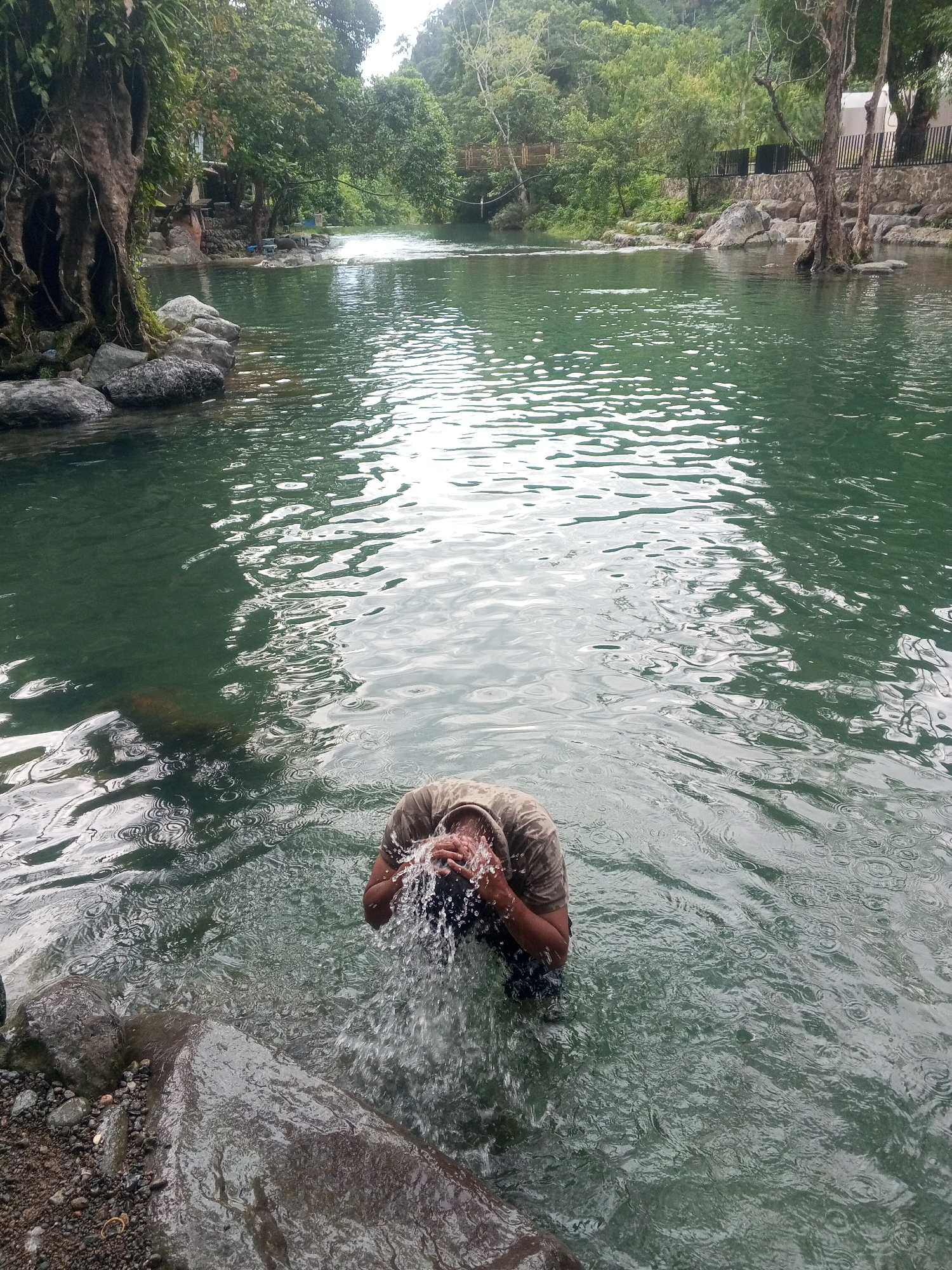 A man is splashing water on his face in a river in Pudeng, Indonesia. He is standing in the water near the rocky bank. The river is wide and surrounded by lush green trees and vegetation. A pedestrian bridge is visible in the background.