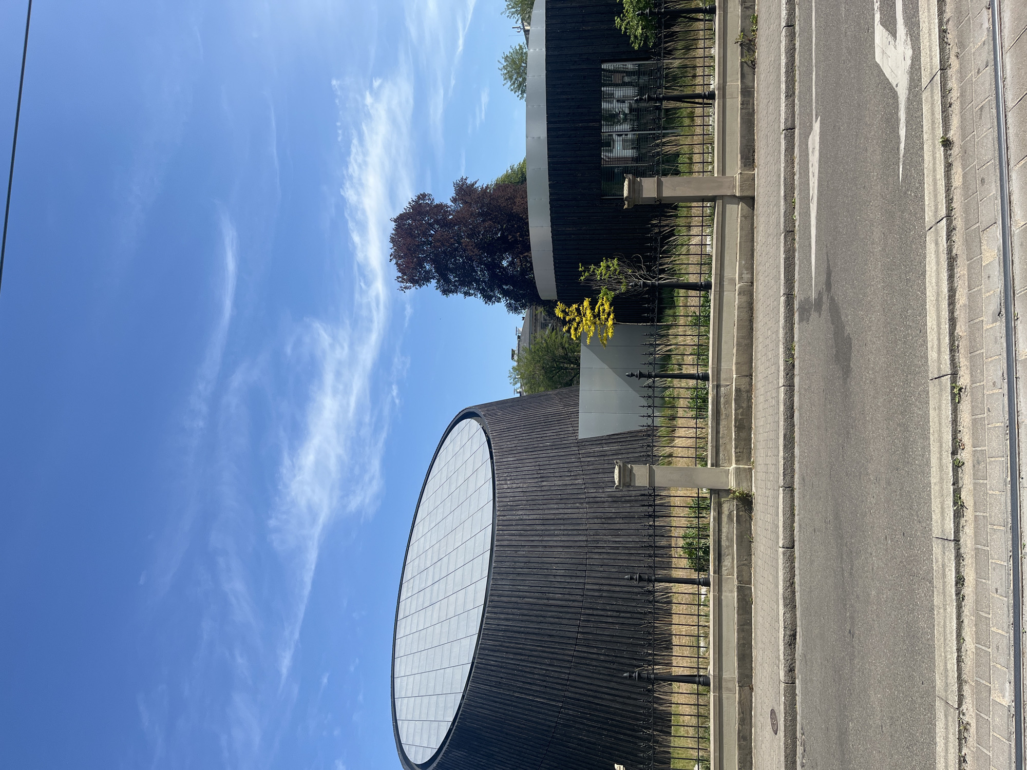 A sunlit urban scene in Strasbourg, France, captures a distinctive modern building under a clear blue sky with wispy clouds. The image is taken during the day, indicated by bright, even lighting.
The most prominent feature is a large, dark circular building on the left, featuring vertical dark siding. A significant portion of its facade is dominated by a huge, round, gridded window or architectural element, reflecting the sky. To the right of this building, partially obscured by a black metal fence, is a large tree with dark, reddish-brown foliage, possibly a copper beech. Further right, another building with a similar architectural style is visible behind more trees and foliage. In front of the buildings and fence, there's a sidewalk and a paved asphalt road. White arrows are painted on the road surface, indicating traffic direction, likely straight ahead. The sidewalk, made of concrete slabs, runs parallel to the road. A black wrought-iron fence separates the landscaped area around the buildings from the public sidewalk and road. Small bushes with yellow-green leaves are also visible near the fence. A dark utility line crosses the upper left corner of the sky. No people or specific activities are visible, presenting a calm and static street view.