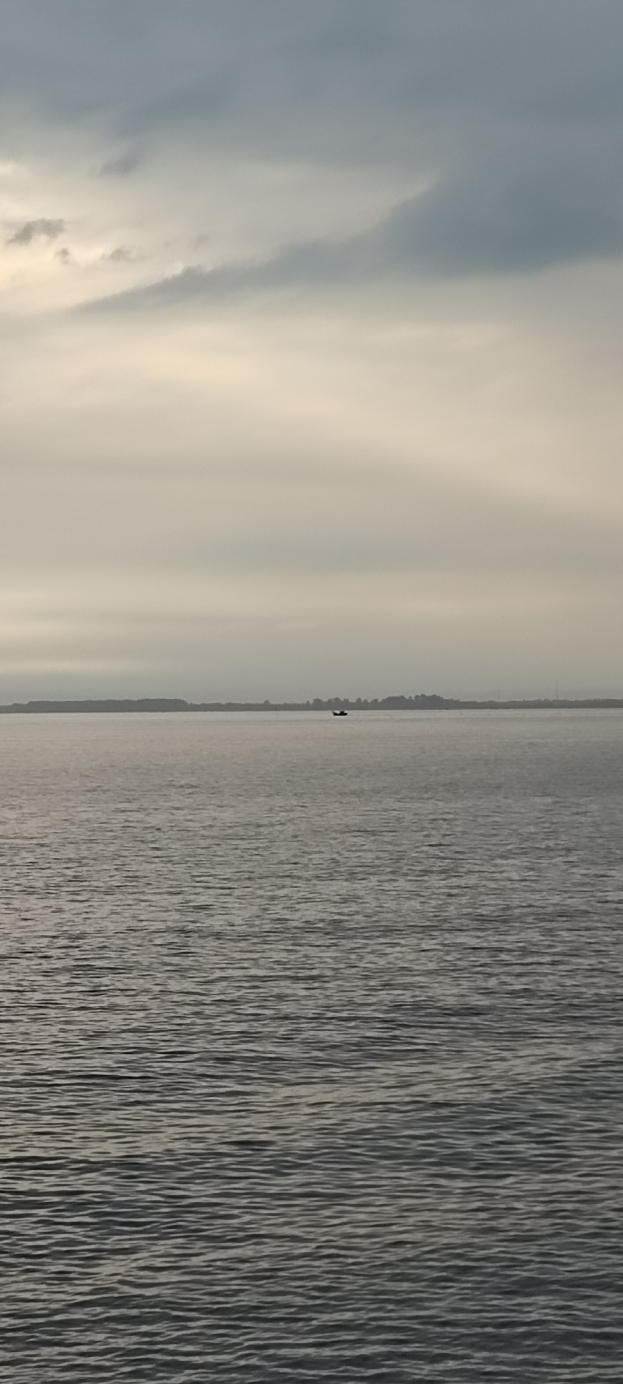 The image depicts a wide, calm body of water, likely a bay or strait, under a cloudy sky. The water exhibits gentle ripples. In the distance, a faint shoreline is visible, characterized by a line of trees or vegetation and possibly some structures. A small, dark boat is present on the water, positioned towards the right of the frame, approximately in the middle third of the image horizontally. The overall atmosphere is subdued, suggesting overcast weather or twilight. Without specific landmarks or identifying features, pinpointing the exact location within Lhokseumawe is not possible from this image alone.