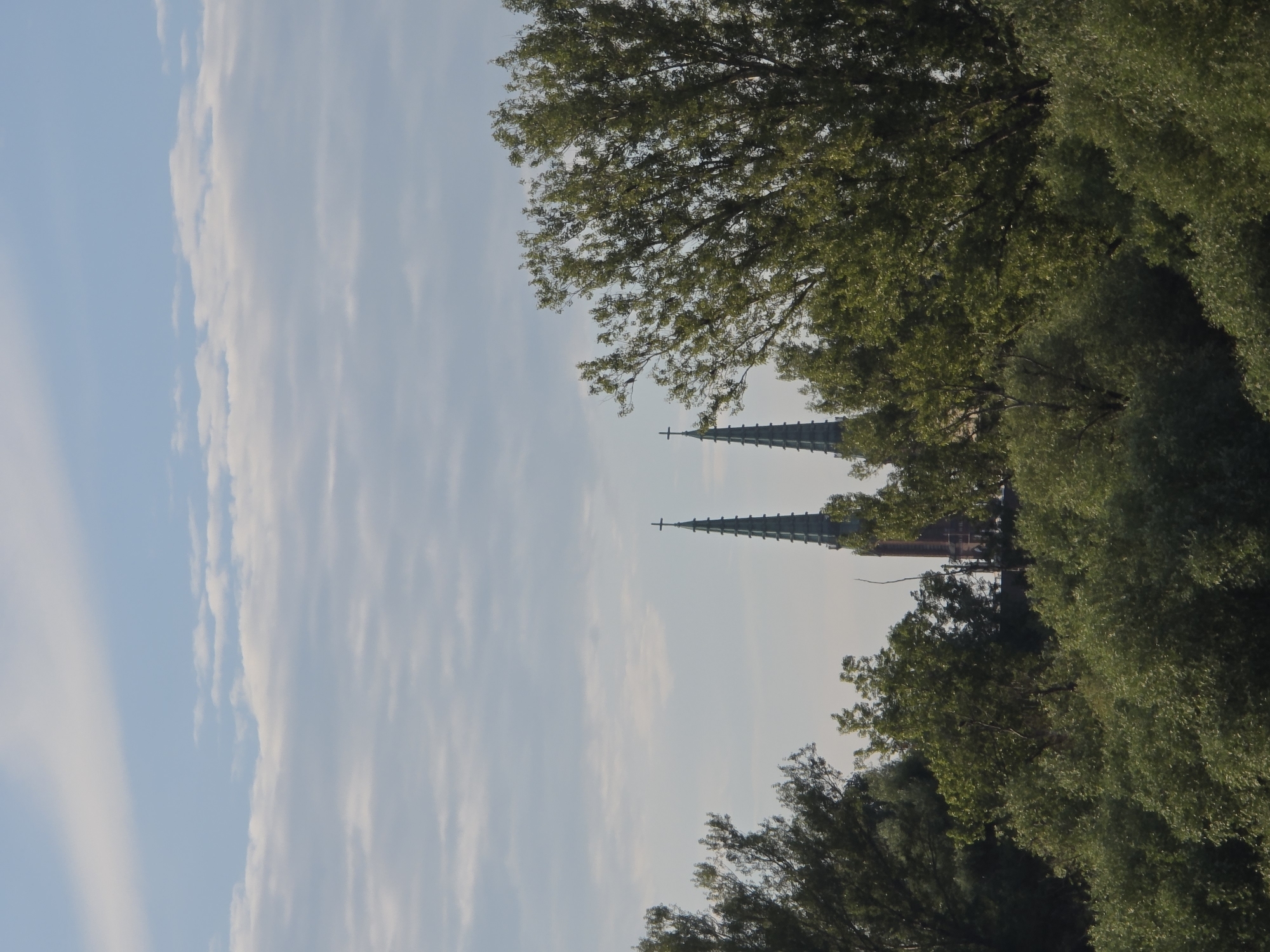 The image shows a view of two church spires, partially obscured by trees, under a cloudy sky. One spire on the left is a dark green, pointed structure topped with a cross. To its right, a similar spire is visible, also topped with a cross. Behind and between the spires, a reddish-brown building with arched windows can be seen, suggesting it is part of the church structure. In the foreground and middle ground, lush green trees with dense foliage dominate the scene, with their branches reaching towards the sky and obscuring parts of the church. The sky is a pale blue with soft, white clouds scattered across it.
The spires are likely those of St. Mary's Cathedral in Warsaw, Poland. The cathedral is a prominent landmark located in the Old Town of Warsaw. The view suggests a perspective from across a body of water, such as the Vistula River, with the trees in the foreground representing vegetation along the riverbanks. The overall atmosphere is serene and somewhat overcast.
