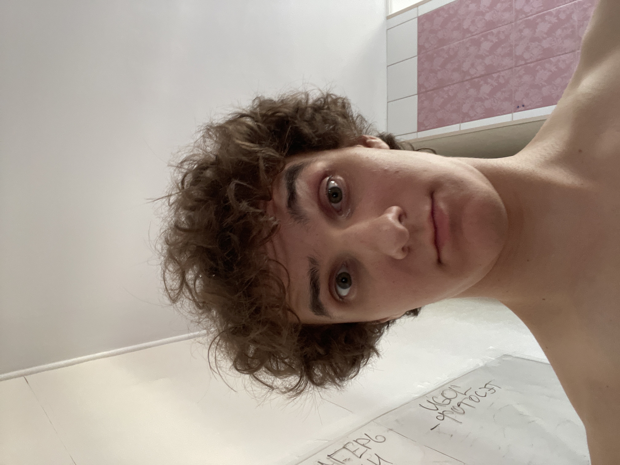 The image is a selfie taken from a low angle, looking up at a young person with curly brown hair. They are looking directly at the camera with wide eyes and a neutral expression. The background consists of a white wall with some writing on it, and a tiled wall with a pink floral pattern. To the left of the frame, there is a vertical white strip, possibly a corner or a doorway. The overall lighting is bright, suggesting an indoor environment. There is no indication of the specific location within Krasnoyarsk, Russia, based on the visual information provided.