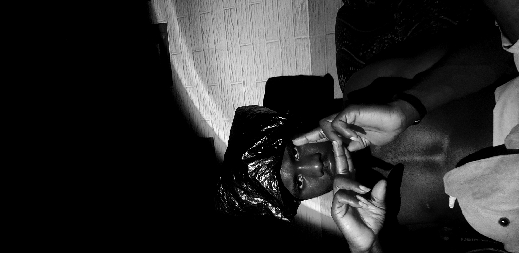 A black and white, medium shot image shows a person wearing a black plastic bag as a head covering. The person is looking directly at the camera with their hands held up in front of their face, fingers intertwined, forming a gesture. The person appears to be in a dimly lit room with a brick wall in the background, illuminated by a light source casting shadows on the wall and the person's face and body. The person's chest and abdomen are visible and appear to be bare. The lower portion of the image shows part of a stuffed toy and some fabric. The overall scene is dark and moody, with dramatic lighting creating strong contrasts. The location is specified as Narayi, Nigeria.