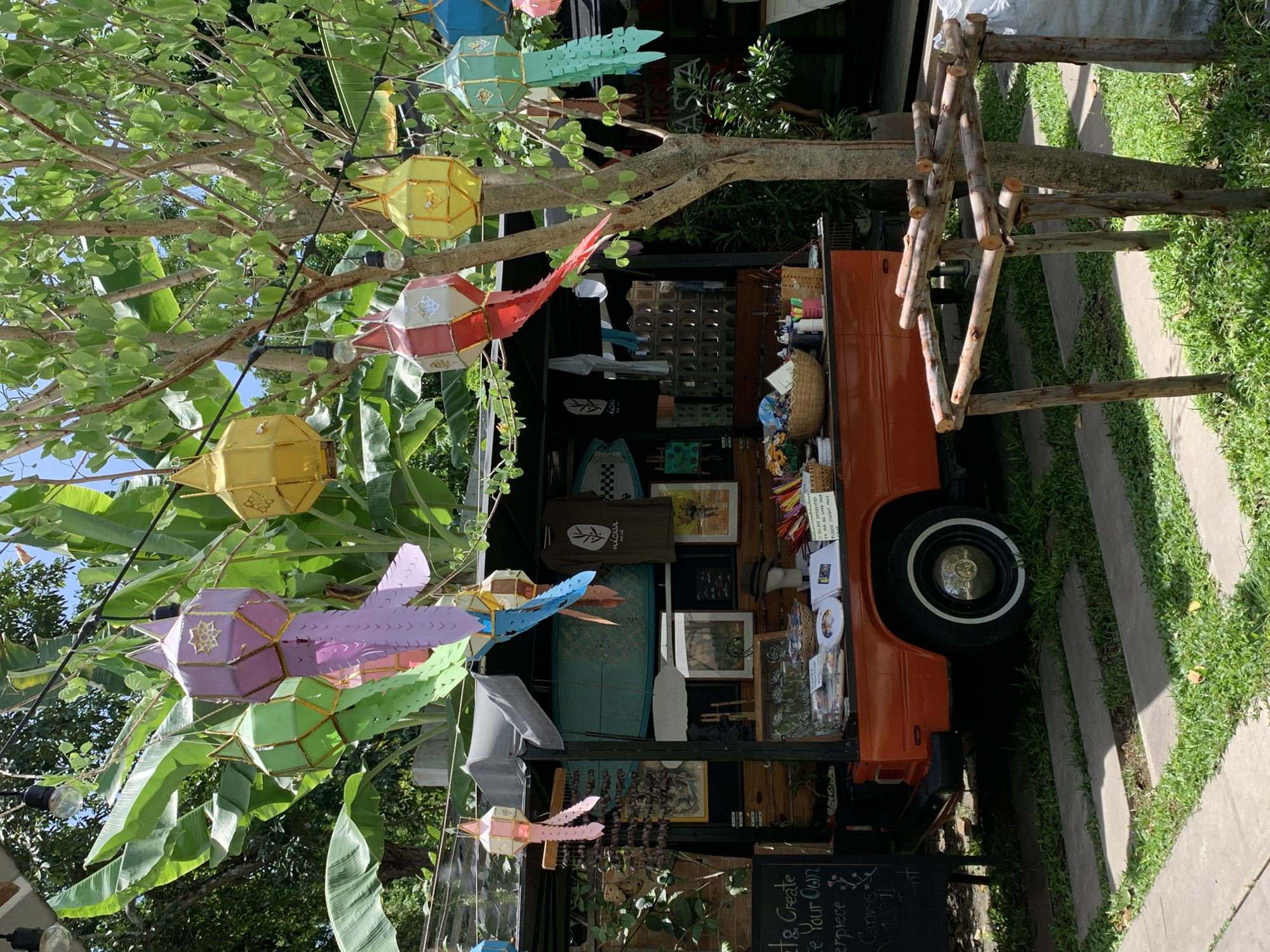 The image depicts an outdoor scene at a market stall, likely in Ko Samui District, Thailand, given the tropical foliage and style of decoration. The primary subject is a vintage orange van converted into a mobile shop. Hanging from strings above the van are colorful, traditional paper lanterns in various shapes and colors, including green, purple, yellow, red, and blue. Behind the van, lush green banana leaves and the branches of a tree provide a natural canopy.
The van's rear is open, revealing shelves stocked with various items. These include what appear to be surfboards, framed artwork or photographs, baskets filled with goods, and art supplies like paints and brushes. A dark-colored blackboard sign to the left of the van displays handwritten text, possibly indicating a workshop or products for sale. In the foreground, a rustic wooden frame constructed from branches stands on a patch of grass. The ground is paved with concrete slabs interspersed with green grass. The overall atmosphere suggests a vibrant, artistic, and relaxed market setting in a tropical location. No people are visible in the frame.