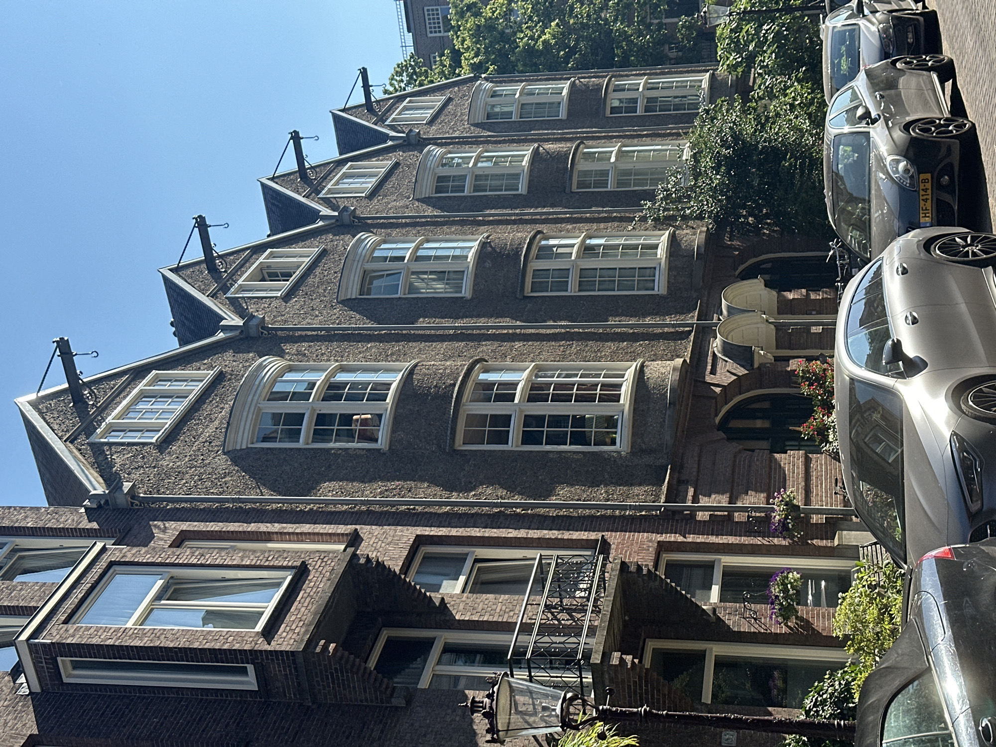 This is a medium-shot, eye-level, outdoor, daytime photograph taken in Amsterdam, Netherlands, on Prinsengracht 185. The scene depicts the facade of a historic brick building with a textured stone overlay. The building has multiple bay windows with white frames and mullions, reflecting the bright blue sky. The roofline features several gabled dormers, each with a single window and decorative iron hooks extending outward.
In the foreground, a dark grey convertible is parked on the cobblestone street to the left, partially obscuring a lighter grey sedan with a license plate reading "HF-414-B". Another dark colored vehicle is parked behind the sedan. Lush green foliage and flowering plants in window boxes are visible on the building's facade. A black wrought-iron lamppost stands to the left of the frame. The overall impression is a sunny day in a charming, historic European city.