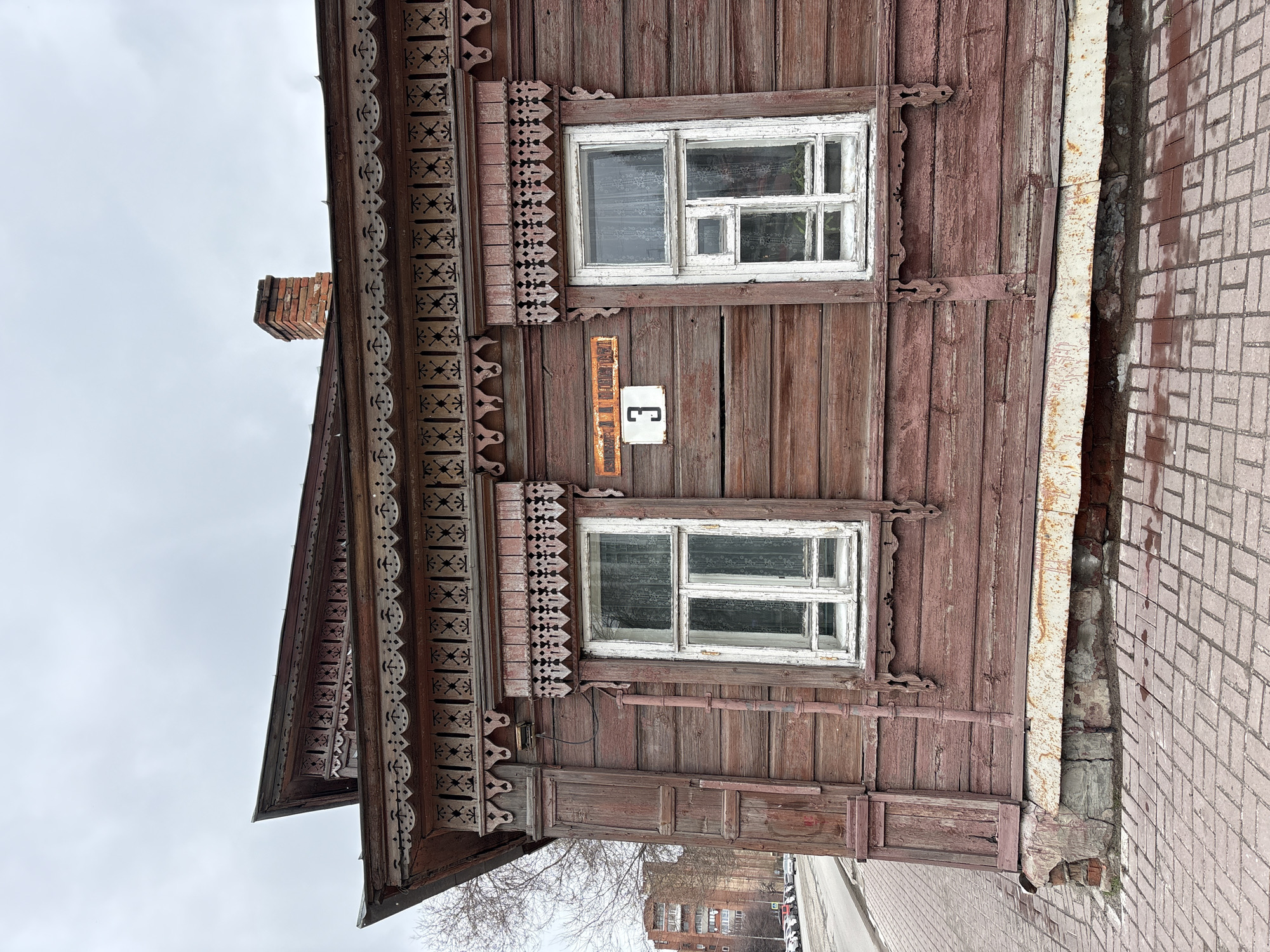 A weathered, single-story wooden house stands in Ulyanovsk, Russia. The house, painted in shades of brown, features two windows with white frames and ornate wooden trim above each. A small square sign with the number "3" is affixed to the wall between the windows. Decorative wooden carvings adorn the eaves of the roof. To the right of the house, a brick sidewalk is visible. A rusty metal pipe runs vertically along the edge of the house where it meets the sidewalk. In the background, a multi-story brick building and parked cars indicate an urban setting. The sky is overcast.