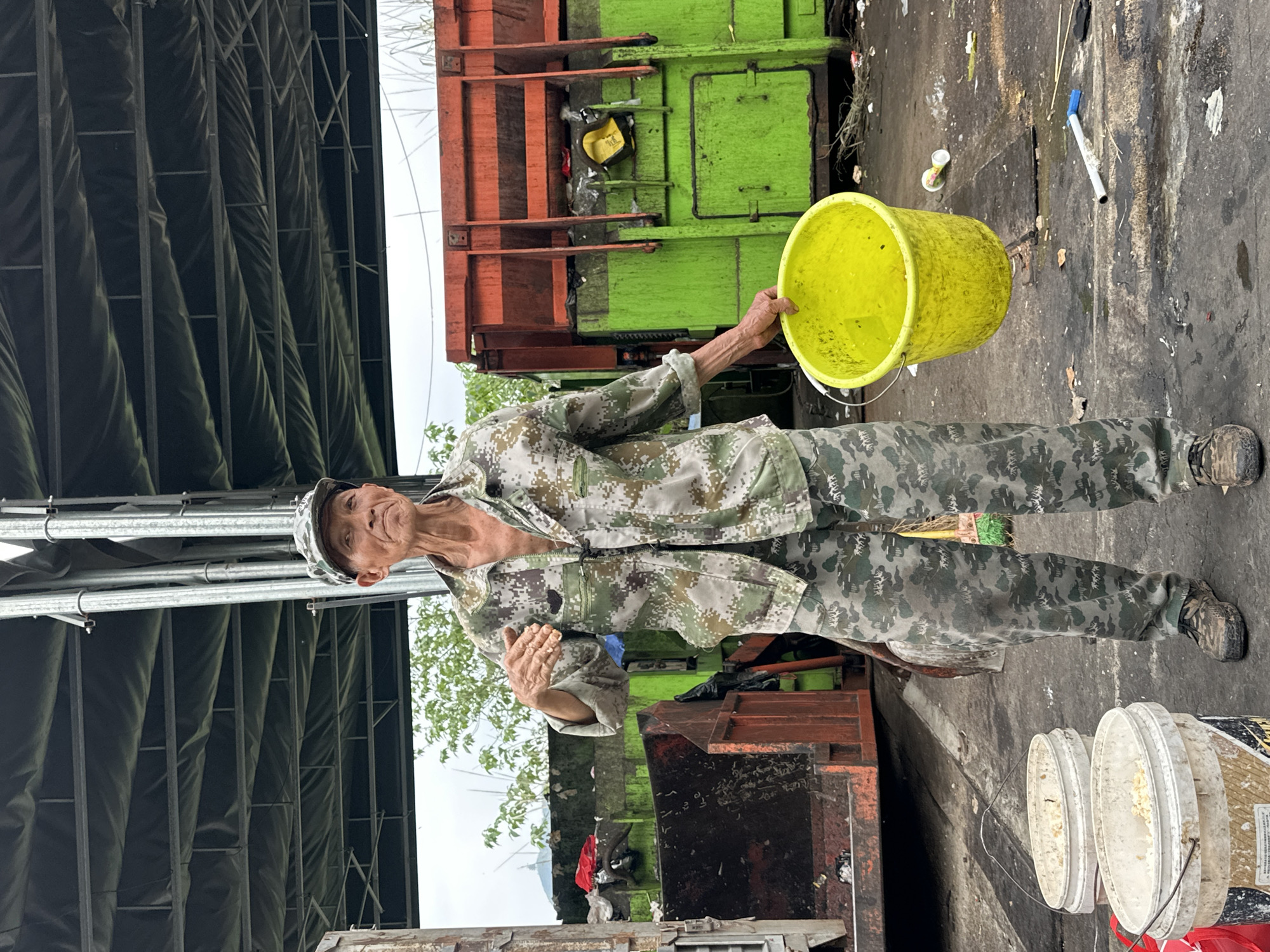 An elderly man, wearing camouflage clothing and a matching hat, stands outdoors holding a yellow bucket. He is positioned near a green and orange garbage truck. The ground is paved and littered with debris, including a pen and several cups. Additional white buckets are visible in the lower right of the frame. The background features what appears to be a structure with black panels and metal supports. The location is Jiangjin, China.