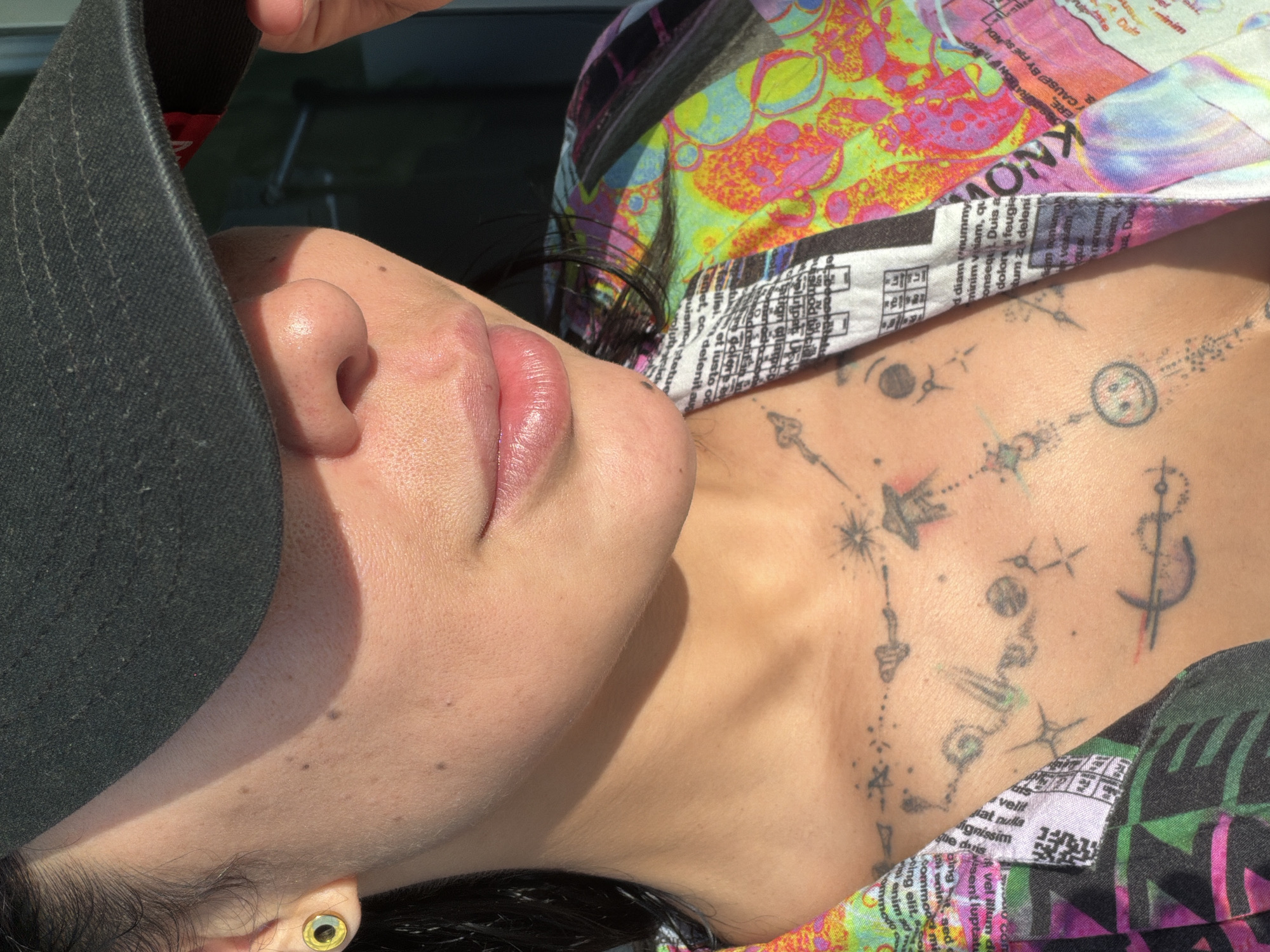 Here is a concise description of the image:
The image shows a person's profile view from the neck up, facing slightly away from the camera. They are wearing a black hat and a brightly colored shirt with text printed on it. The person has tattoos on their neck and chest. The location is not discernible from the image, so information about L'Hospitalet De Llobregat, Spain cannot be determined from the image provided.