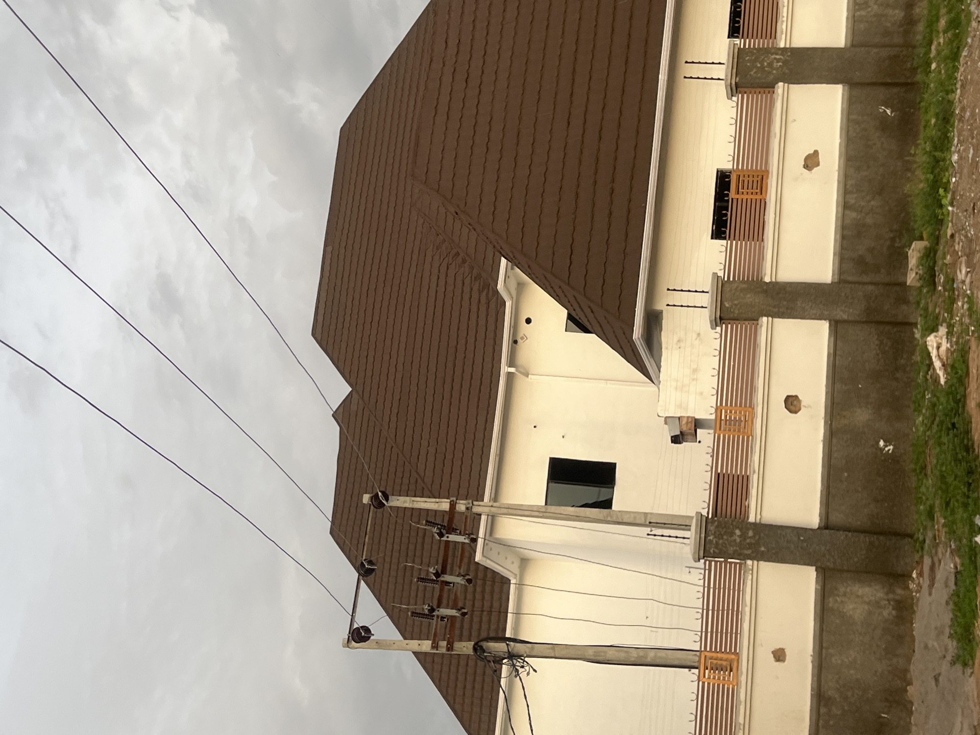 The image shows the exterior of a building in Howayi, Nigeria. The building has a brown tiled roof and light-colored walls with brown decorative elements. Windows are visible on the side of the building. A utility pole with electrical lines runs along the side of the building. The sky is overcast. Vegetation can be seen on the right side of the image.