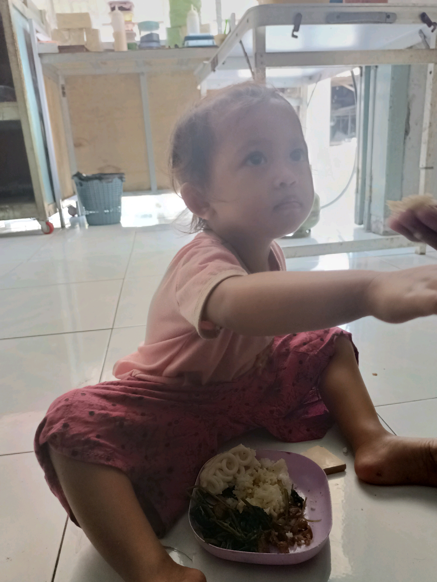 A young child, possibly a toddler, sits on a tiled floor in what appears to be a domestic setting. The child is wearing a pink t-shirt and red patterned shorts. Their body is turned to the right, with their right arm extended towards the viewer. A small plate containing rice and some green vegetables is placed to the child's right. In the background, there are shelves with various items, a blue trash can, and open doorways leading to another area. The lighting suggests it is daytime, with sunlight filtering into the room. The floor is tiled in a light color. The overall environment suggests a simple living space.