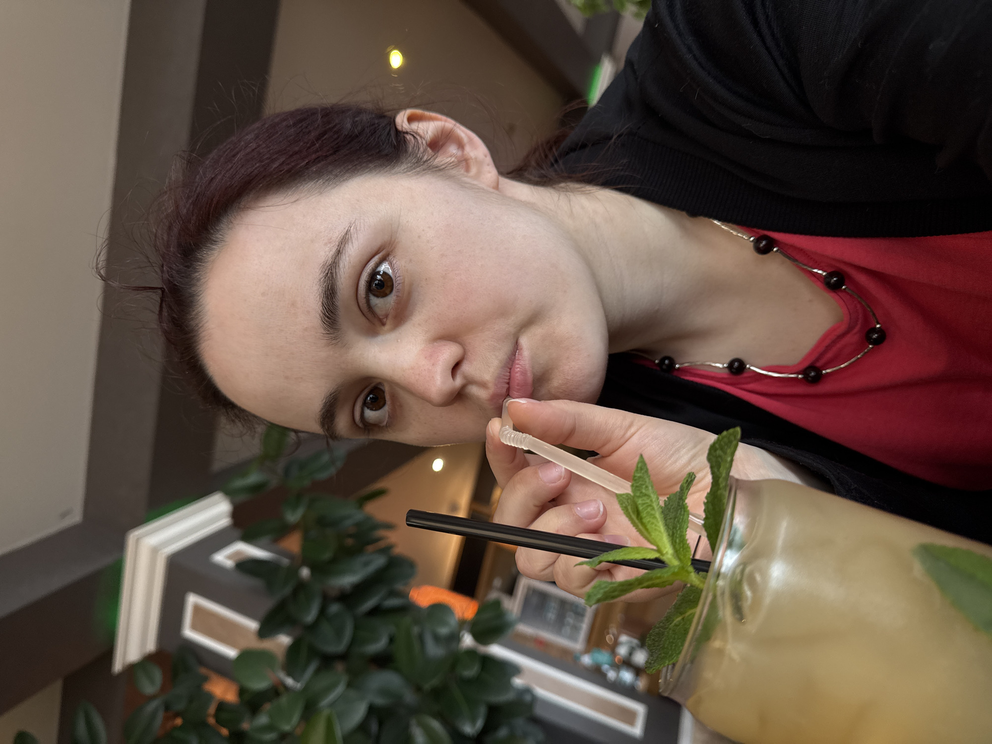A woman is holding a drinking glass with a beverage in it. The glass also contains two drinking straws, one black and one white. The glass is decorated with sprigs of mint leaves. The woman is holding the white straw up to her lips. She is wearing a red shirt underneath a black cardigan and a necklace made of silver and black beads. She has brown hair and brown eyes. The background appears to be a restaurant or cafe, with a green plant in the foreground. The location cannot be determined from the image.