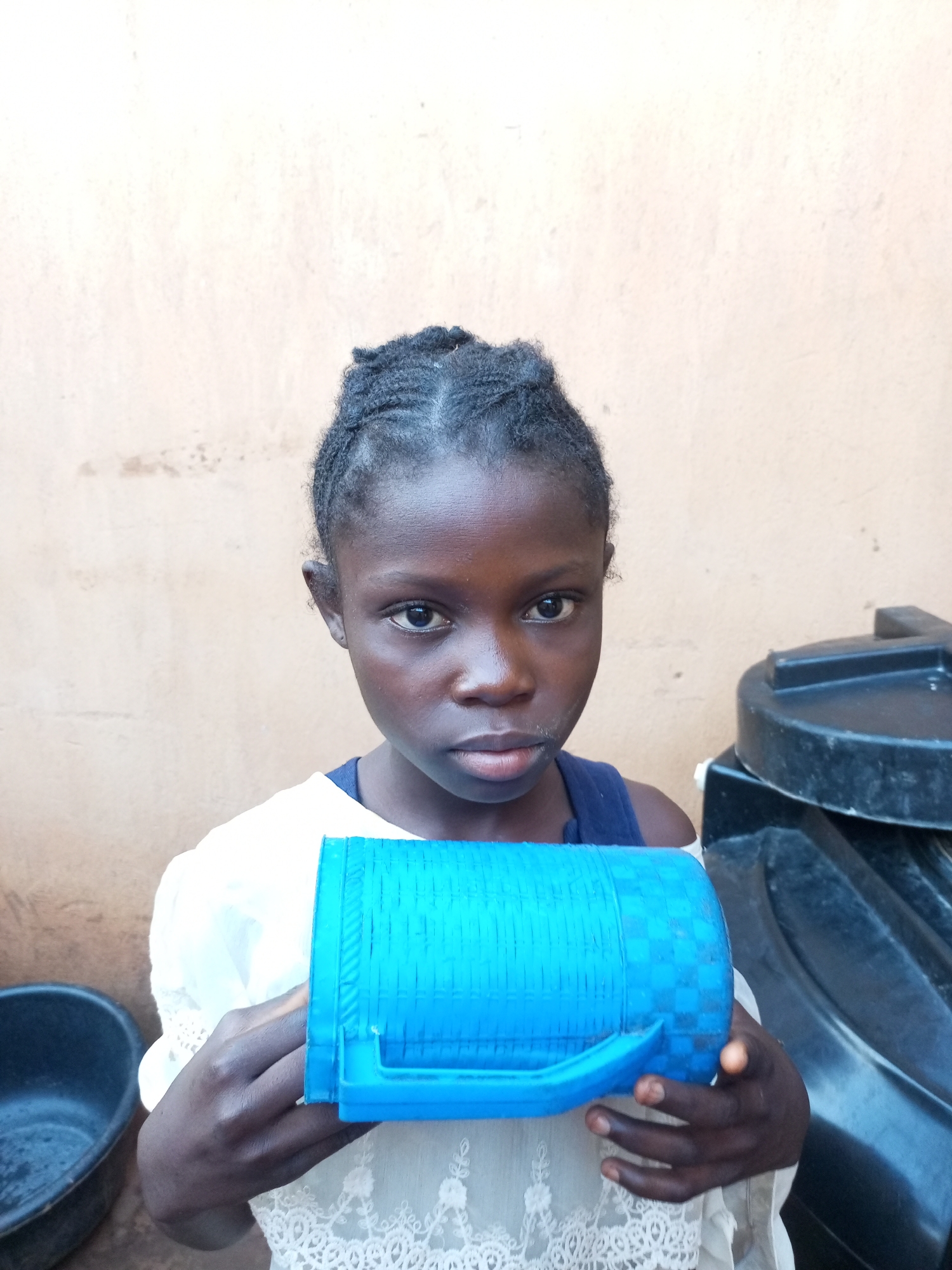 A young Black girl with dark skin and braided hair is holding a blue plastic mug in front of her. She is wearing a white top with lace trim and a dark blue undershirt. The girl is looking directly at the camera with a serious expression. Behind her is a light brown wall. To her right, a black plastic water tank is partially visible. To her left, a black basin is also visible. The scene appears to be an outdoor setting in Tempure, Nigeria.