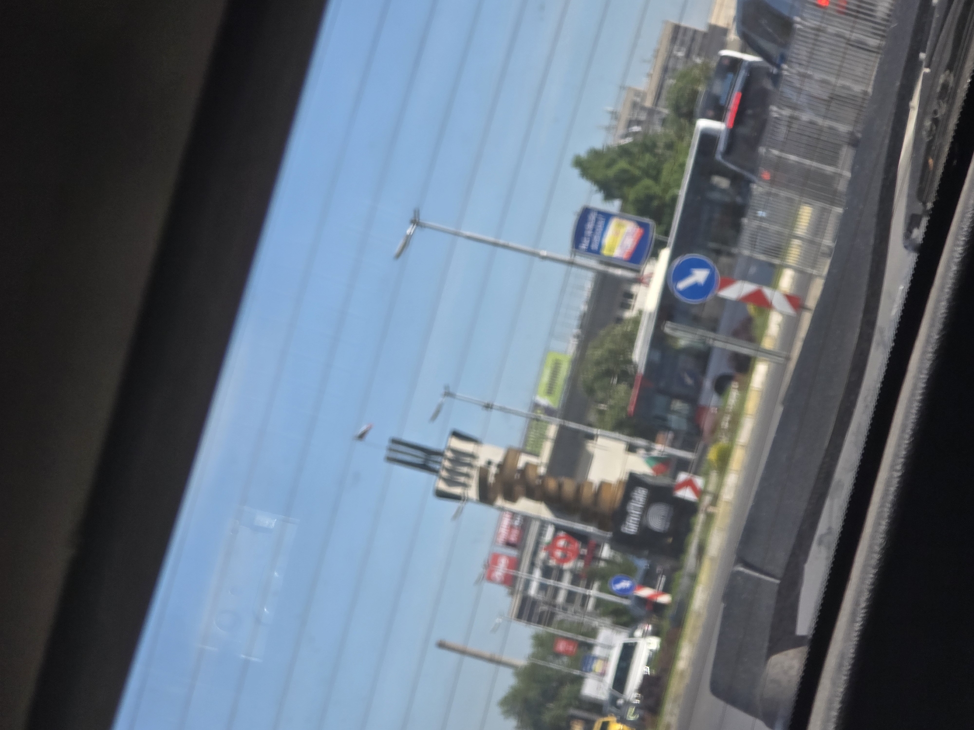 The image shows a view through a vehicle window, possibly a car. Outside the window, the scene depicts a busy street in an urban environment. There are multiple signs visible, including traffic signs and advertisements. A bus is present in the traffic flow. The environment suggests a city location, but the specific location is not identifiable from the information given.