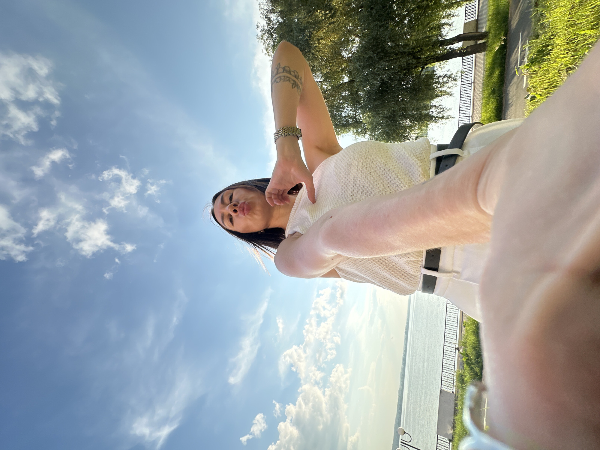 A low-angle, wide-shot, outdoor selfie shows a young woman in a white top and white pants with a black belt, posing with puckered lips and her arm extended toward the camera, a tattoo visible on her forearm. She is positioned in the foreground of a sunny scene with a bright blue sky dotted with white clouds. Behind her, a calm body of water, likely a reservoir or lake, stretches towards the horizon, bordered by a paved walkway, lush green grass, and trees. Streetlights with multiple globes are visible along the walkway on the left side of the image. The location is implied to be a waterfront area within Votkinsk, Russia, likely along the Kama River or Votkinsk Reservoir, given the presence of the wide expanse of water and the waterfront promenade. The sunlight suggests it is daytime, possibly late afternoon or early evening due to the warm glow on the foliage.