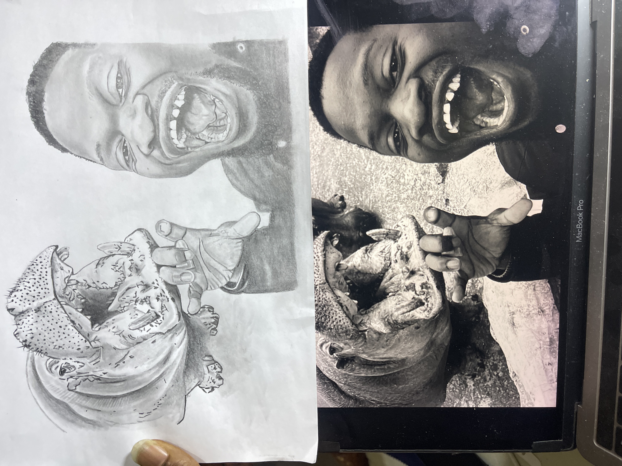 The image displays a physical pencil drawing held above a MacBook Pro laptop. The drawing, executed in grayscale on white paper, depicts a hippopotamus with its mouth wide open, exposing teeth and tongue, interacting with a smiling man whose mouth is also wide open in laughter, showing his teeth and tongue. The man's right hand, wearing a ring on the middle finger, is positioned as if holding the hippopotamus's lower jaw. A partial human hand with light brown skin is visible on the far left, holding the drawing. Below the drawing, a MacBook Pro laptop is visible, displaying a grayscale photograph that is the apparent source material for the drawing, showing the same hippopotamus and laughing man composition. The laptop's screen shows the lower half of the photograph. The laptop itself is black with visible "MacBook Pro" text on the bottom bezel, and minor dust particles are visible on its surface. No visual cues within the image indicate a specific location in Jos, Nigeria.