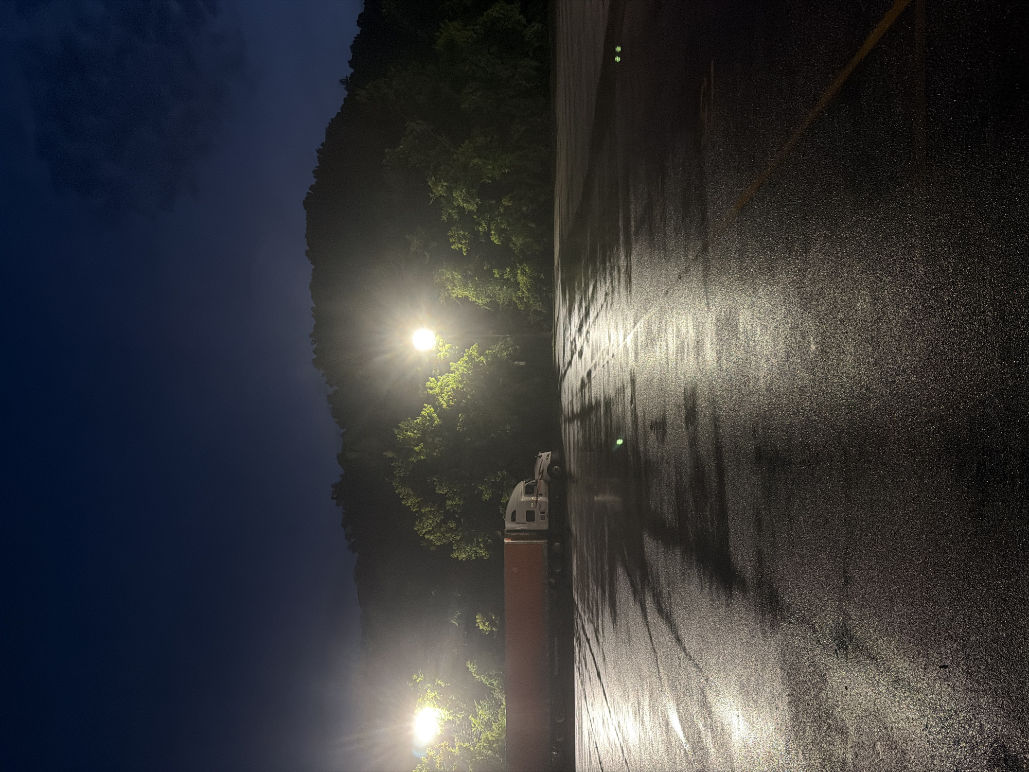 A semi-trailer truck is parked in a wet, empty parking lot at night. Two tall light poles illuminate the scene, casting long reflections on the dark, wet asphalt. Trees and a dimly lit hillside form the background. The sky is a deep blue, transitioning to black at the top of the frame. The truck is white and orange, with the trailer appearing to be a deep red or orange color. No people are visible in the image. The overall impression is one of stillness and isolation. The location is an outdoor parking lot in Wildwood, United States.
