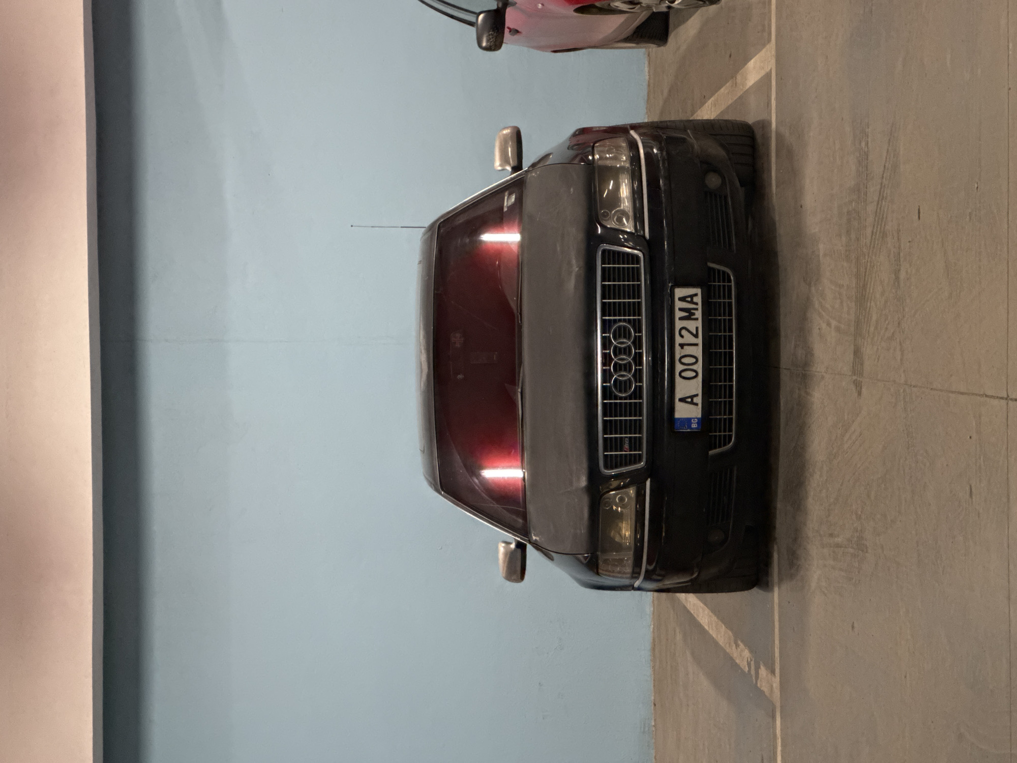 A black Audi car, with license plate "A 0012 MA", is parked in an indoor parking garage. The car is facing the camera. The background consists of a blue wall on the left and a concrete floor and a wall on the right. Another vehicle, partially visible, is parked in front of the Audi. The scene appears to be dimly lit. The location is unspecified, but the license plate suggests the car is located in Bulgaria.