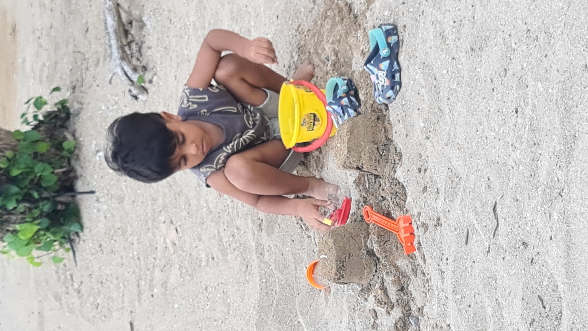 The image depicts a young child playing in the sand on a beach, likely in Denpasar, Indonesia. The child, with dark hair, is wearing a patterned sleeveless shirt and shorts, and is crouching down, engrossed in building sandcastles. In front of the child are two sand structures, a yellow bucket with a red handle, an orange toy rake, and a small red toy boat. A pair of dark blue Crocs with an ice cream cone pattern are placed on the sand to the right of the child. The beach appears to be a mix of sand and small pebbles, with some vegetation visible in the background near a tree trunk. The lighting suggests it is daytime.